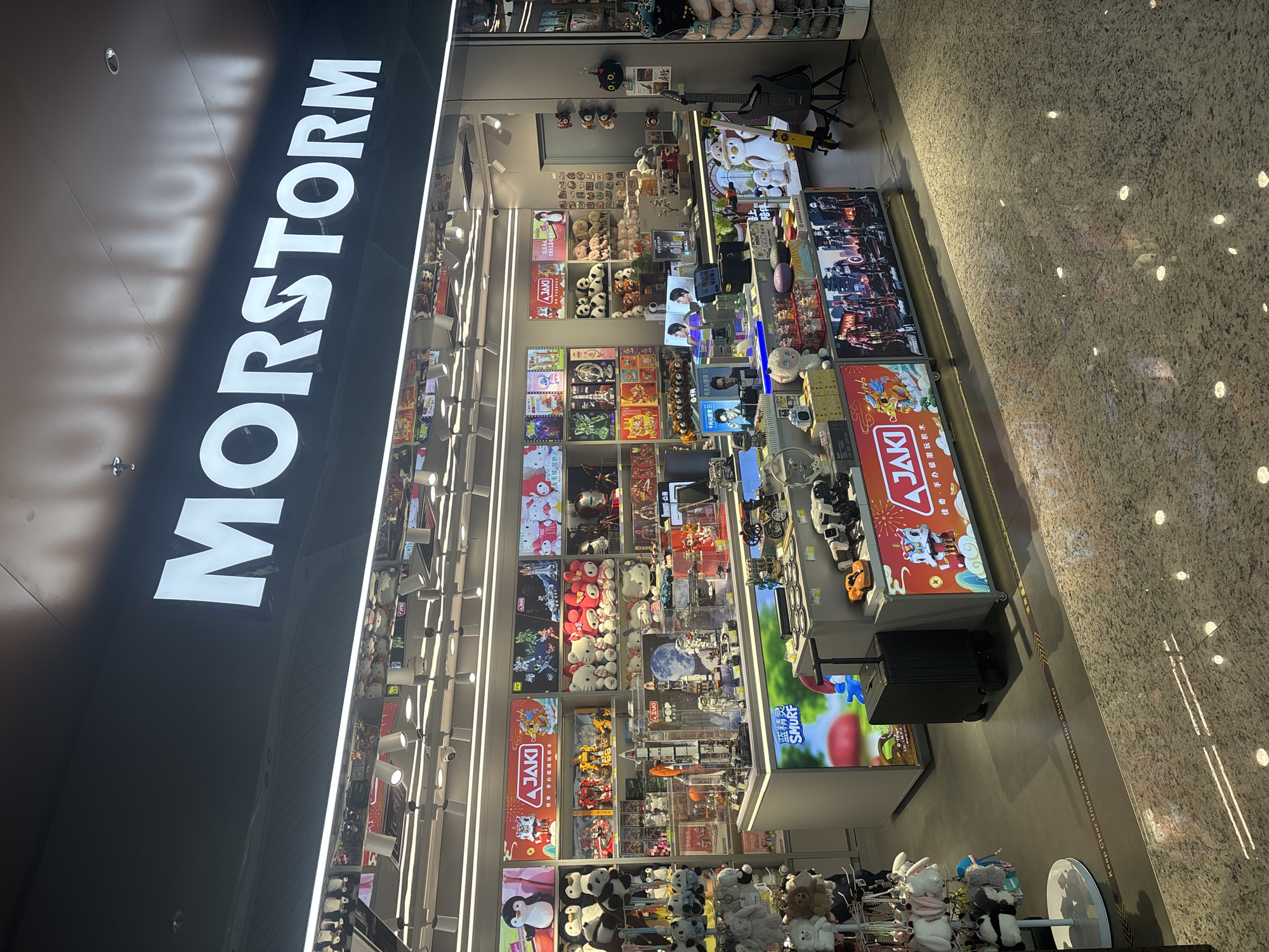 The image shows the storefront of a store named "MORSTORM" located in Baiyun District, China. The store appears to be a toy or collectible shop. The facade is black with large white illuminated letters spelling out "MORSTORM". Below the sign, the store interior is visible, filled with shelves stocked with various items. These include plush toys, action figures, model kits, and other merchandise. There are display cases showcasing some of the products, and a large digital display on the front counter shows images of characters. To the left of the entrance, there is a display of keychain plush toys. A black rolling suitcase is parked near the entrance. The floor is tiled with a speckled pattern, and there are spotlights illuminating the store. The overall impression is that of a well-lit retail space selling consumer goods.