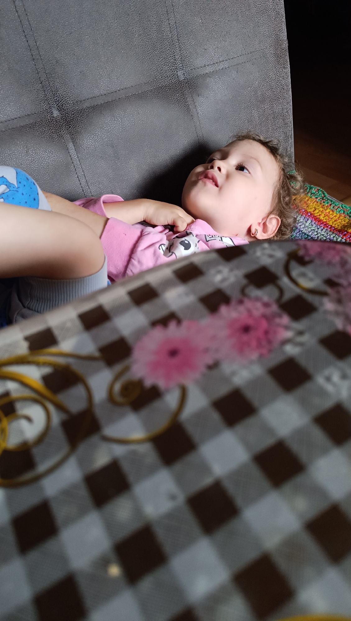 A young child with curly brown hair and wearing a pink shirt is lying on a gray sofa, looking upwards. The child has on a pair of light blue shorts with a cartoon character print. In front of the child is a table covered with a checkered tablecloth that has a floral pattern. The background is dark, with a sliver of natural light falling on the sofa and the child's face. The image appears to be taken indoors, in a domestic setting. There is no indication of the child's location being in the city of Karsha, Russia.