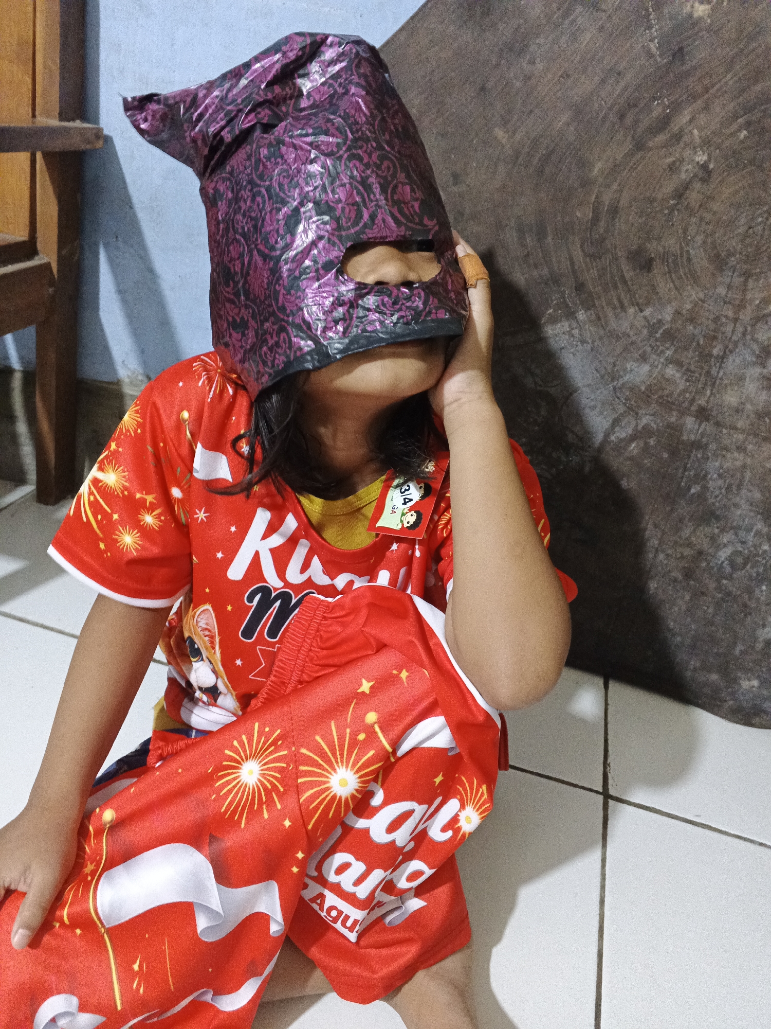 A child, wearing a red shirt with a yellow collar and red pants adorned with fireworks, sits on a tiled floor. The child's head is covered by a purple, patterned bag that has been fashioned into a mask, with cutouts for the eyes. The child's right hand is touching the mask. The background includes a wooden surface, a wooden chair, and a light blue wall. The scene appears to be indoors, possibly in a home located in Warungkiara, Indonesia.