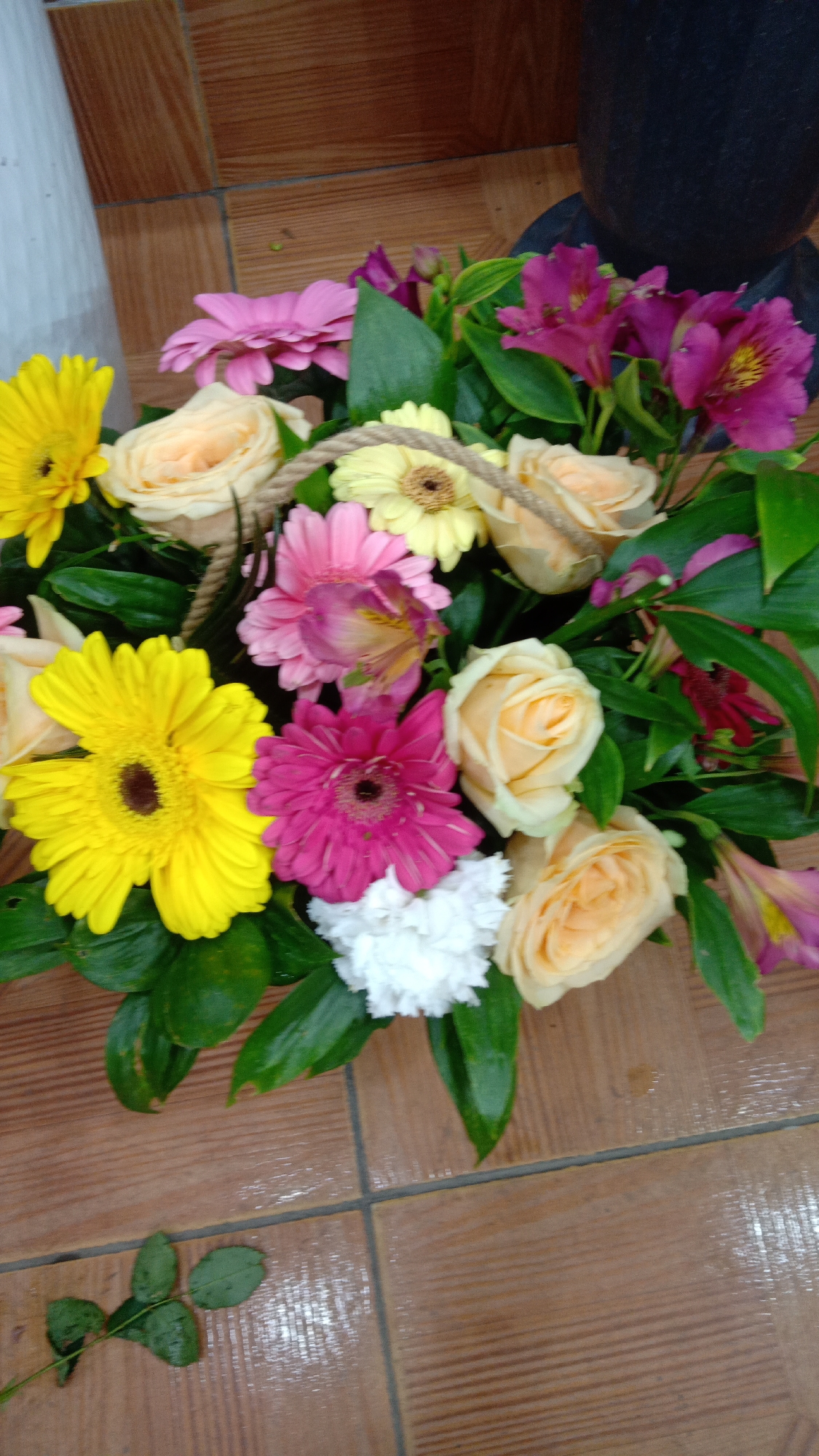 A high-angle medium shot reveals a colorful bouquet of flowers arranged in a basket on a brown tiled surface. The flowers include yellow gerbera daisies, pink and peach roses, pink gerberas, white carnations, and pink alstroemeria. The arrangement is surrounded by green foliage. A stray rose leaf lies on the tiled surface. The scene likely takes place indoors. No people are visible. There is no indication of the location being in Yerevan, Armenia.