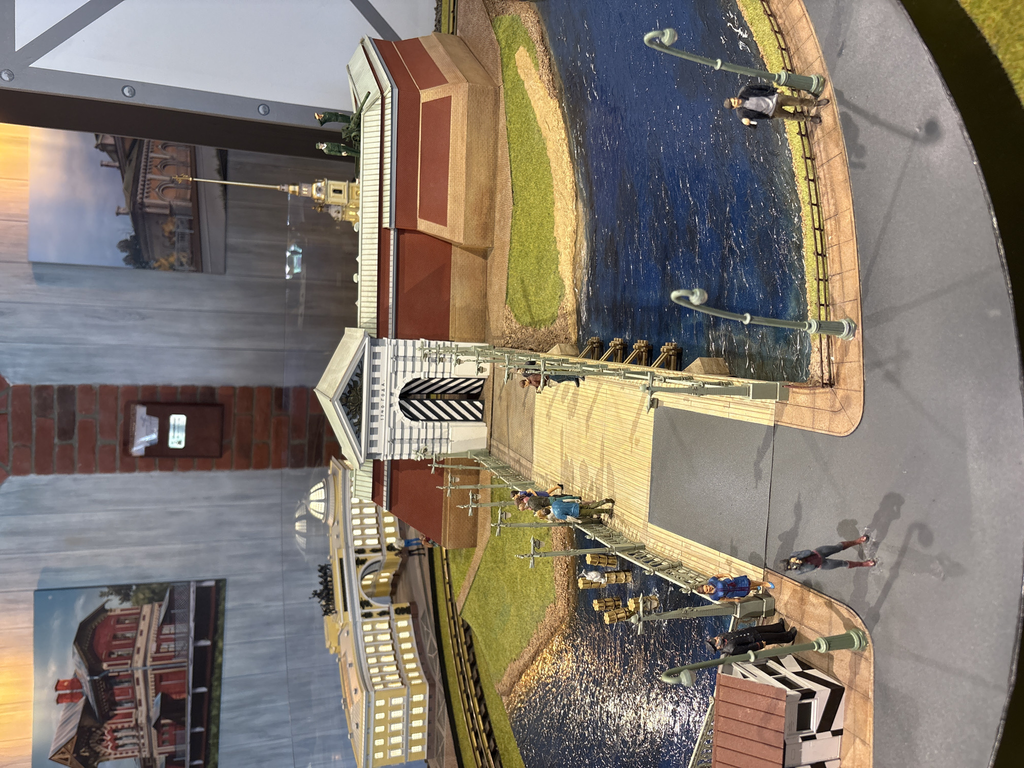 The image shows a miniature model of the Peter and Paul Fortress in Saint Petersburg, Russia. The model includes the fortress itself, a bridge leading to it, a body of water representing the Neva River, and several miniature figures of people and objects.
The Peter and Paul Fortress is depicted with its characteristic red brick walls and a tall, golden spire. The bridge is made of wood and has railings and lampposts. The water is blue and textured to represent waves.
There are several miniature figures in the scene. On the bridge, there are figures of people walking and standing. Near the fortress, there are figures of soldiers and what appears to be a cannon. On the land surrounding the fortress, there is a small patch of green grass.
The background of the image shows a wall with wooden panels and two framed pictures of buildings. One picture on the left shows a red and white building with a steep roof. The picture on the right shows a lighter colored building with arches.
The overall scene is a detailed and realistic depiction of a historical landmark, likely part of a museum exhibit or a diorama.