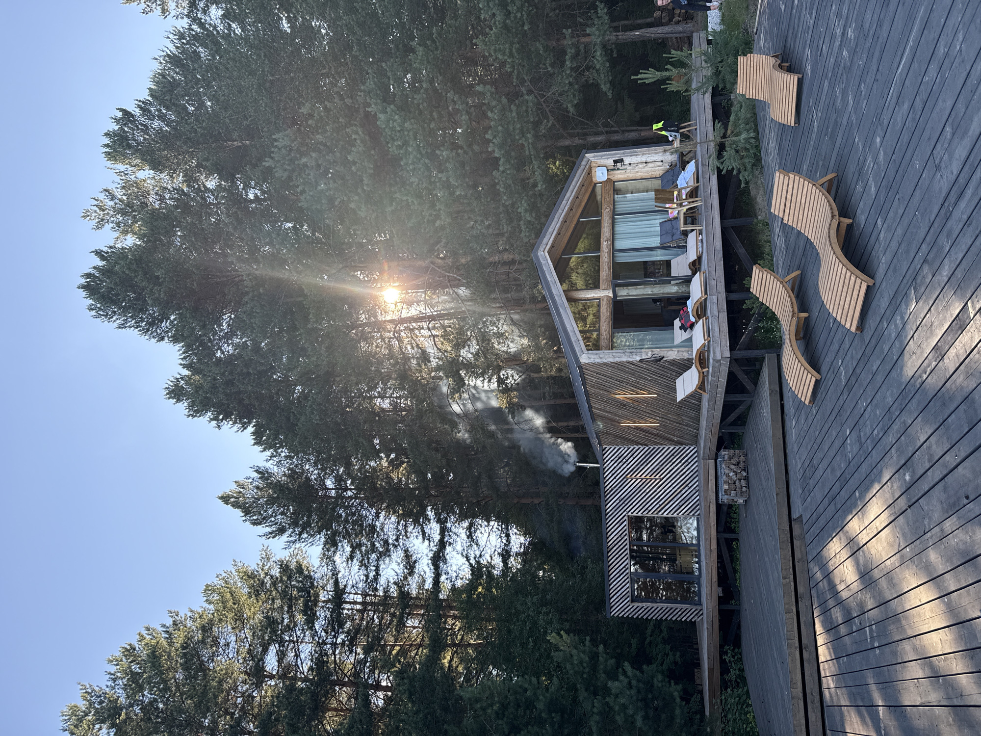 The image depicts a modern wooden house with a large glass facade, nestled within a dense forest of tall pine trees. The house features a slanted roof and wooden siding with horizontal stripes. Smoke is rising from a chimney on the left side of the house. A wooden deck surrounds the house, and several wooden lounge chairs are arranged on the deck. Sunlight streams through the trees, creating lens flares and dappled light on the deck. The environment appears to be a serene, natural setting. The location is specified as Votkinsk, Russia. There are at least three individuals visible in the image: one person standing on the far right edge of the deck near some greenery, and at least two people on the deck of the house, one of whom appears to be sitting.