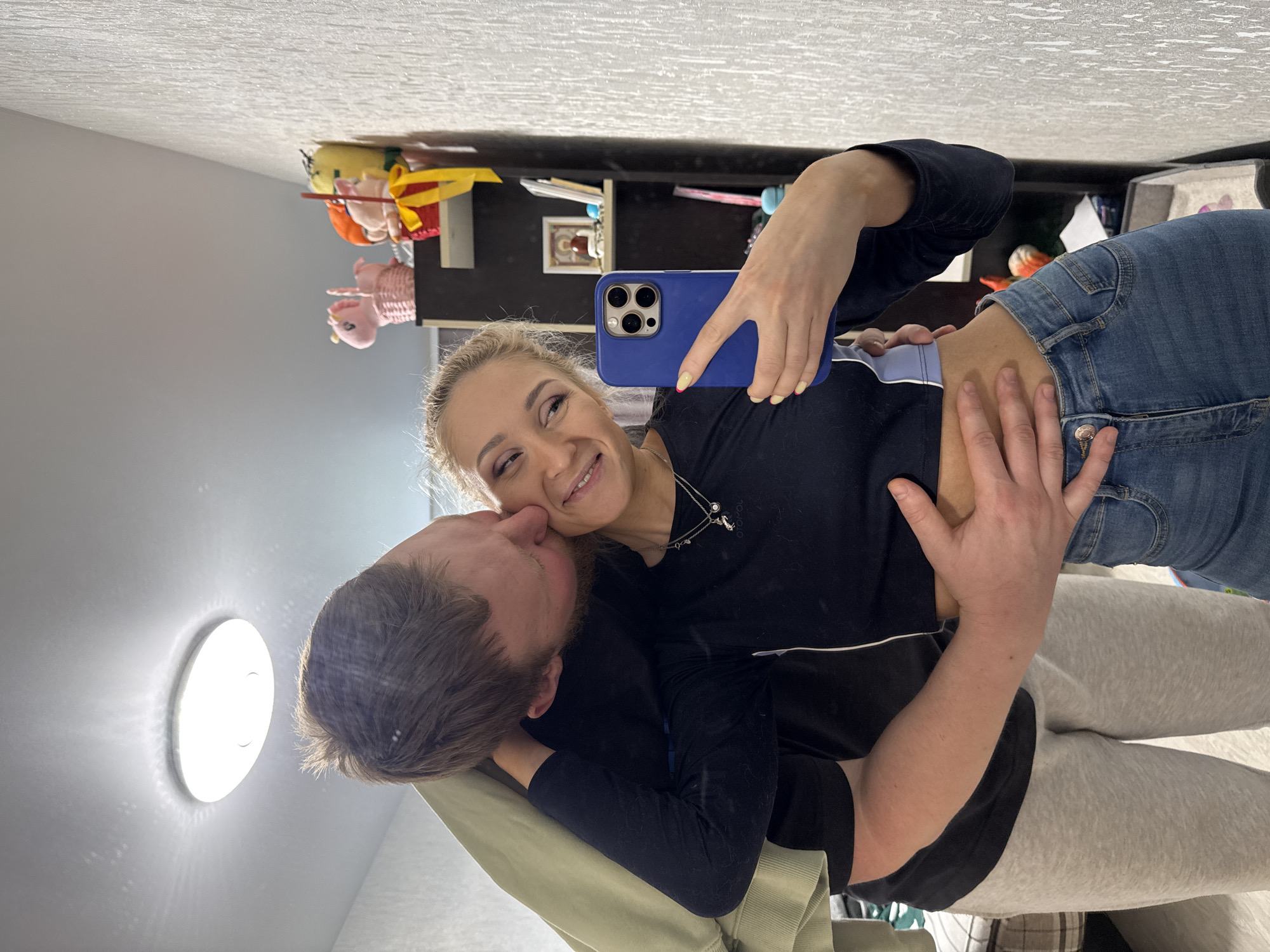 A young couple is posing for a selfie indoors. The woman, who is blonde, holds a blue smartphone and smiles. A man is kissing her cheek. They are both wearing casual clothing. The scene is in a room with a ceiling light, and a shelf containing toys and other items is visible in the background. The location is Vereshchagino, Russia.