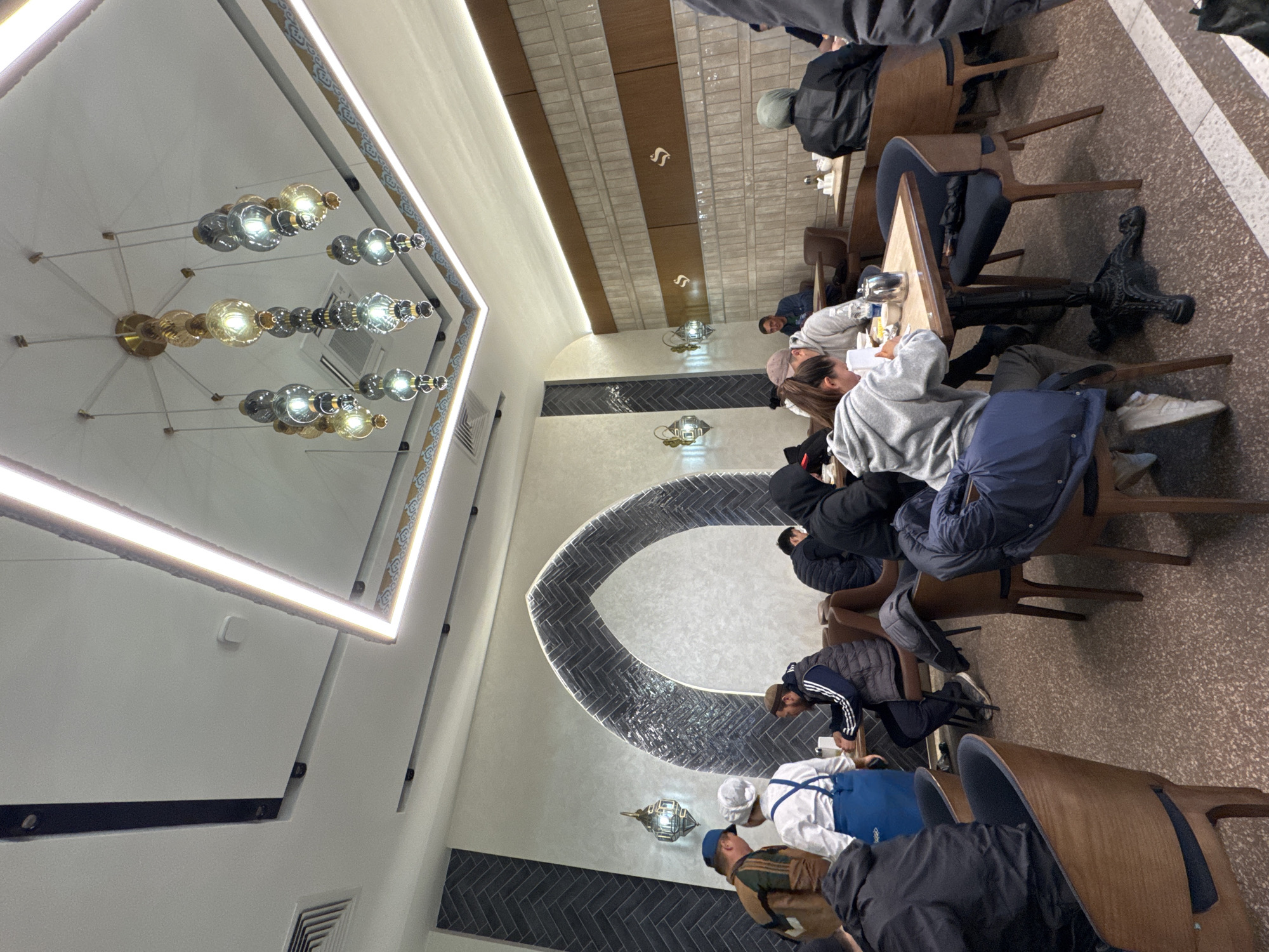 The image captures the interior of a contemporary restaurant or cafe in Bishkek City, Kyrgyzstan, filled with patrons. The scene is oriented vertically, showing the ceiling with ornate lighting fixtures on the left and the dining area with tables and people on the right.
Approximately a dozen individuals are visible, seated at various tables. Most are casually dressed in jackets, hoodies, and long-sleeved shirts, engaged in activities such as dining, looking at phones, or conversing. One person near the foreground is seen wearing a white chef's hat and a blue apron, suggesting they are staff. The overall atmosphere appears relaxed and social.
The establishment features a distinctive interior design. The ceiling is adorned with multiple elaborate chandeliers composed of clusters of gold and silver-toned spherical glass orbs, alongside long, linear LED light strips integrated into hexagonal ceiling panels. The walls exhibit a mix of textures and patterns: some are light-colored, while others feature dark grey or black herringbone tile patterns. A prominent architectural feature is a large, arched section of herringbone tiles creating a decorative focal point on a wall. Upper wall sections incorporate brown and white rectangular tiles with small, stylized white 'S' symbols. Decorative wall sconces with metallic frames and glass elements are also present. The floor is a speckled, light brown material, with some lighter striped patterns visible.
The lighting fixtures are all illuminated, suggesting it might be evening or night, or simply a well-lit indoor space. The location cues, particularly the decorative tile work and lighting, imply a modern yet culturally influenced aesthetic. No specific text is largely visible beyond the stylized 'S' symbols on the wall.
