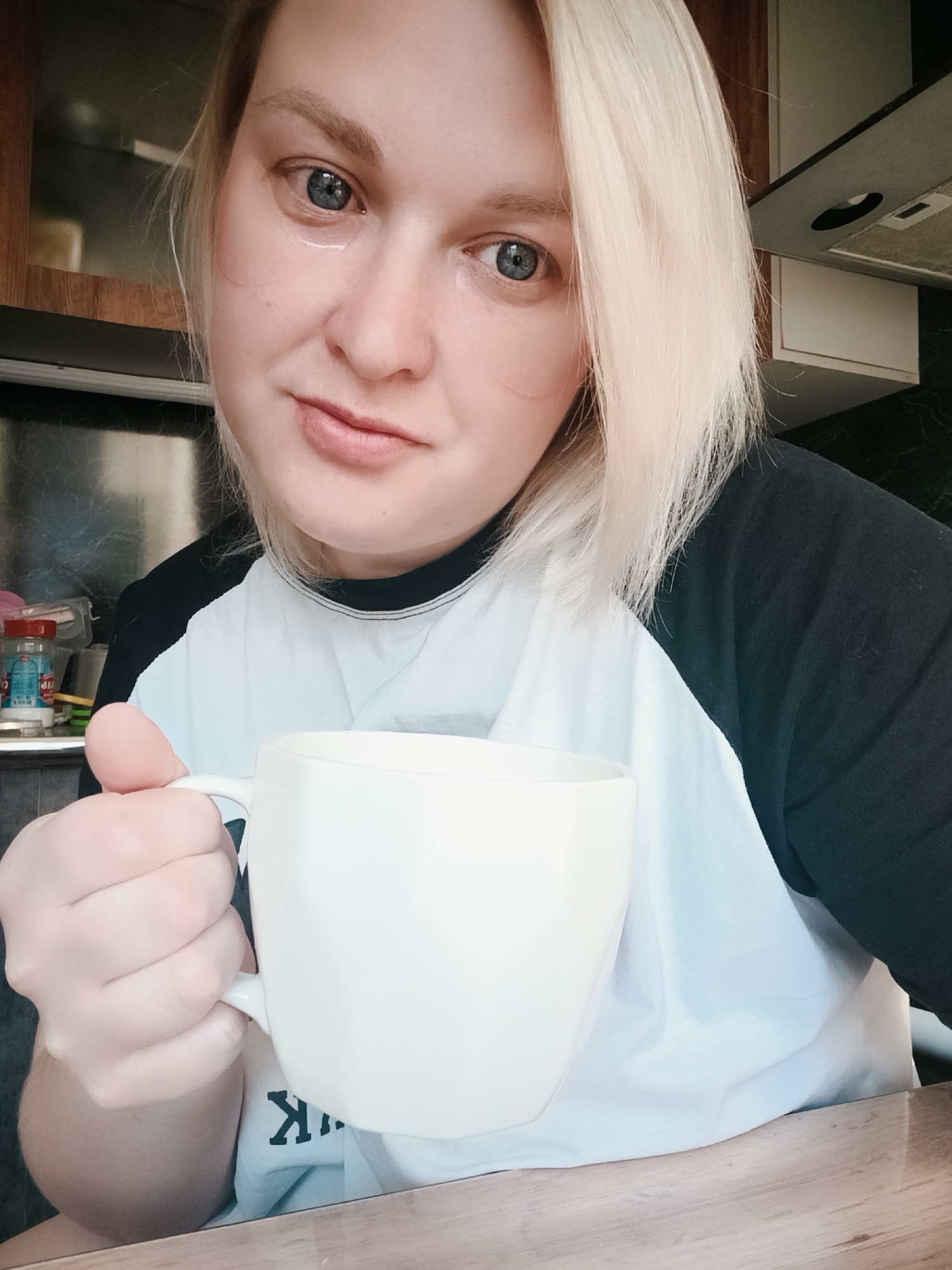 A fair-skinned woman with blue eyes and blonde hair holds a white mug in her right hand. She wears a white t-shirt with black sleeves. She appears to be inside a kitchen, with a wooden countertop in the foreground and kitchen cabinets and appliances visible in the background. Based on image metadata, the image was taken in the city of Chaikovsky, Russia.