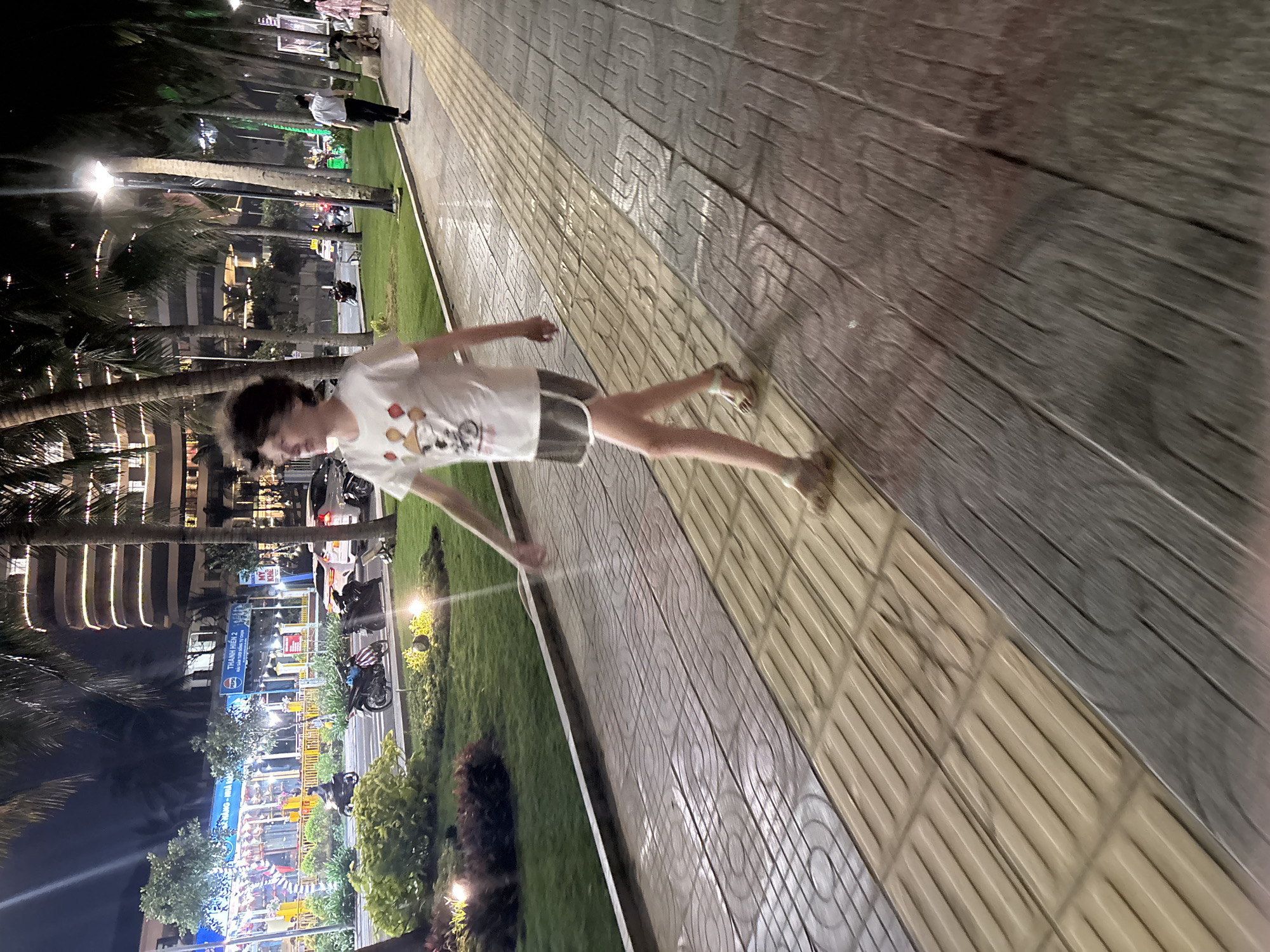 A young person wearing a white t-shirt with cartoon graphics and gray shorts is walking on a two-toned paved sidewalk next to a strip of grass, seemingly at night. The lighting is mixed with artificial illumination from street lights, building lights, and lights on a nearby boardwalk or shopping area. Palm trees are visible along the street. In the background, there are shops and restaurants that could possibly be identified as being located at the Bien Dong Park in Đà Nẵng, Vietnam.