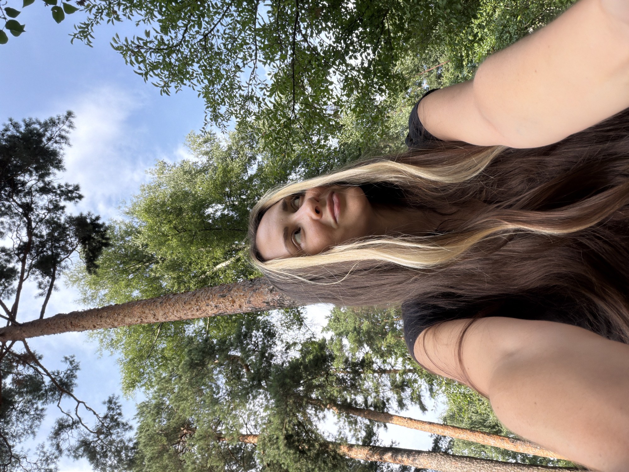 In Udelnaya, Russia, a young woman with long, dark brown hair featuring blonde highlights is captured in a selfie taken outdoors. She is wearing a black t-shirt and is positioned in the foreground, looking up and to her right with a slight smile. The background is dominated by lush green trees and the sky, with tall pine trees prominently visible. The image appears to be taken from a low angle, emphasizing the height of the trees and the open sky above. The lighting suggests it is daytime, with natural sunlight filtering through the foliage. The overall environment is that of a park or forest.