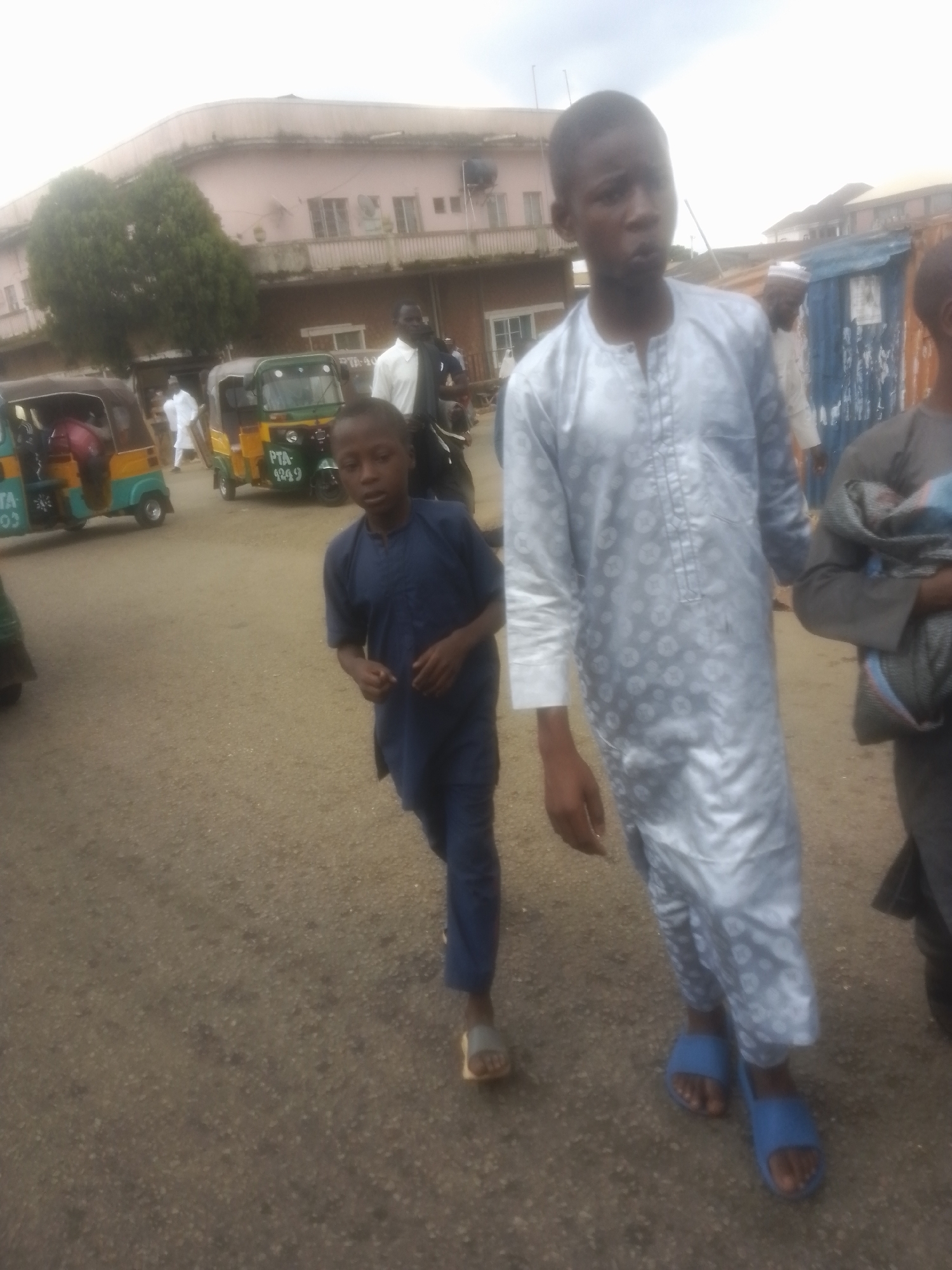 The image depicts a street scene in Jos, Nigeria, likely a bustling market or thoroughfare. In the foreground, two young boys are walking. The boy on the right, taller and closer to the camera, is wearing a silver, patterned tunic and blue slides. The boy on the left, smaller and to his left, is wearing a dark blue tunic and sandals. Behind them, other individuals and vehicles are visible. A yellow and green tricycle, a common mode of transport in Nigeria, is to the left, and another similar vehicle is further back. Buildings line the street, one with a light pink facade and balconies. The ground is paved with asphalt, showing signs of wear. The overall atmosphere suggests a typical day in a Nigerian urban environment.