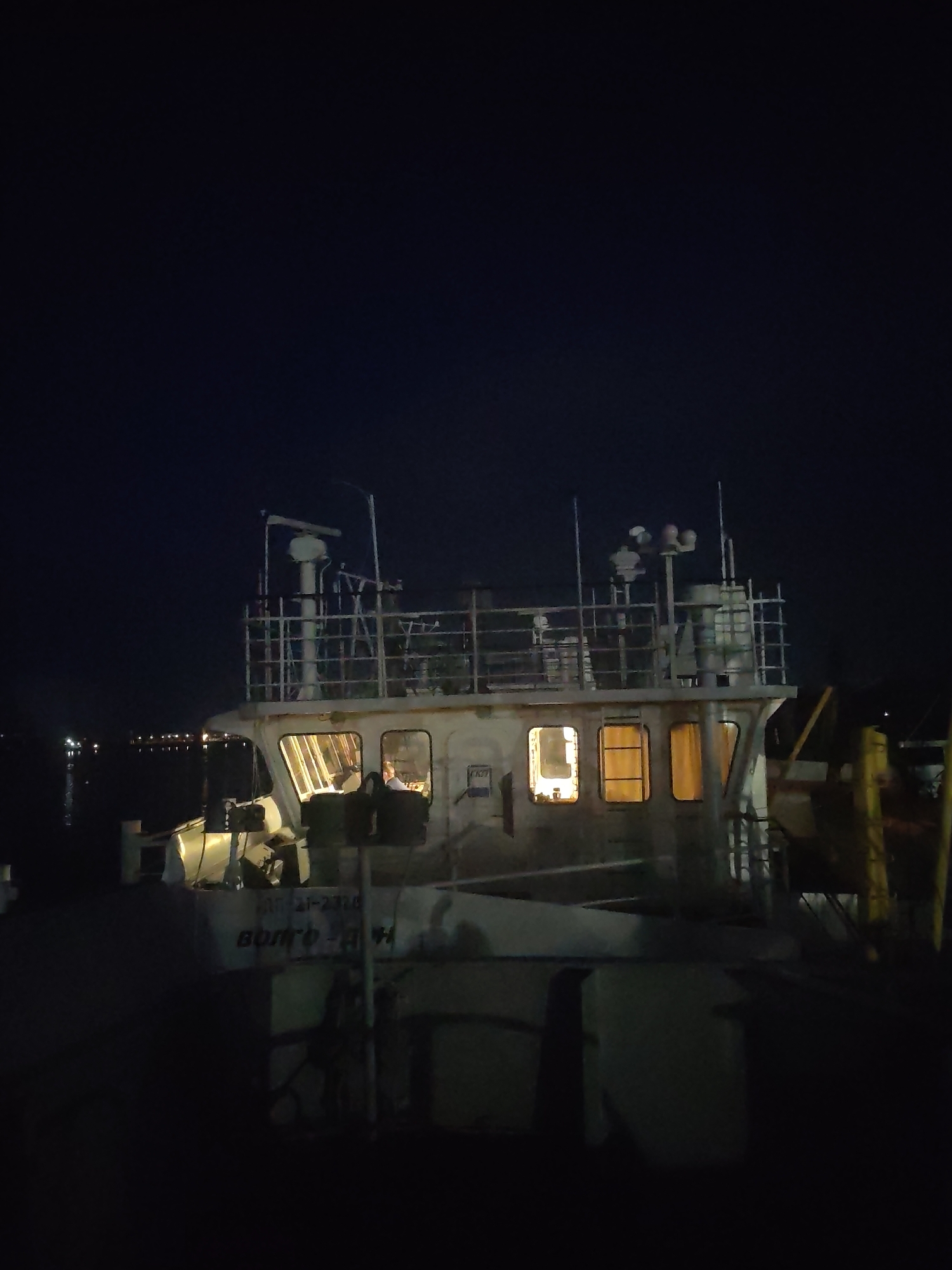 The image captures a vessel at night in Bol'shie Mkhi, Russia. The cabin windows of the boat are illuminated. The top deck of the boat is outlined by a metal railing. Masts with navigational equipment and lights are visible. The name of the boat, “ВОЛГО-ДОН,” is visible on the hull. The background shows distant lights across the water. The rest of the image is dark, suggesting a very dim ambient light.