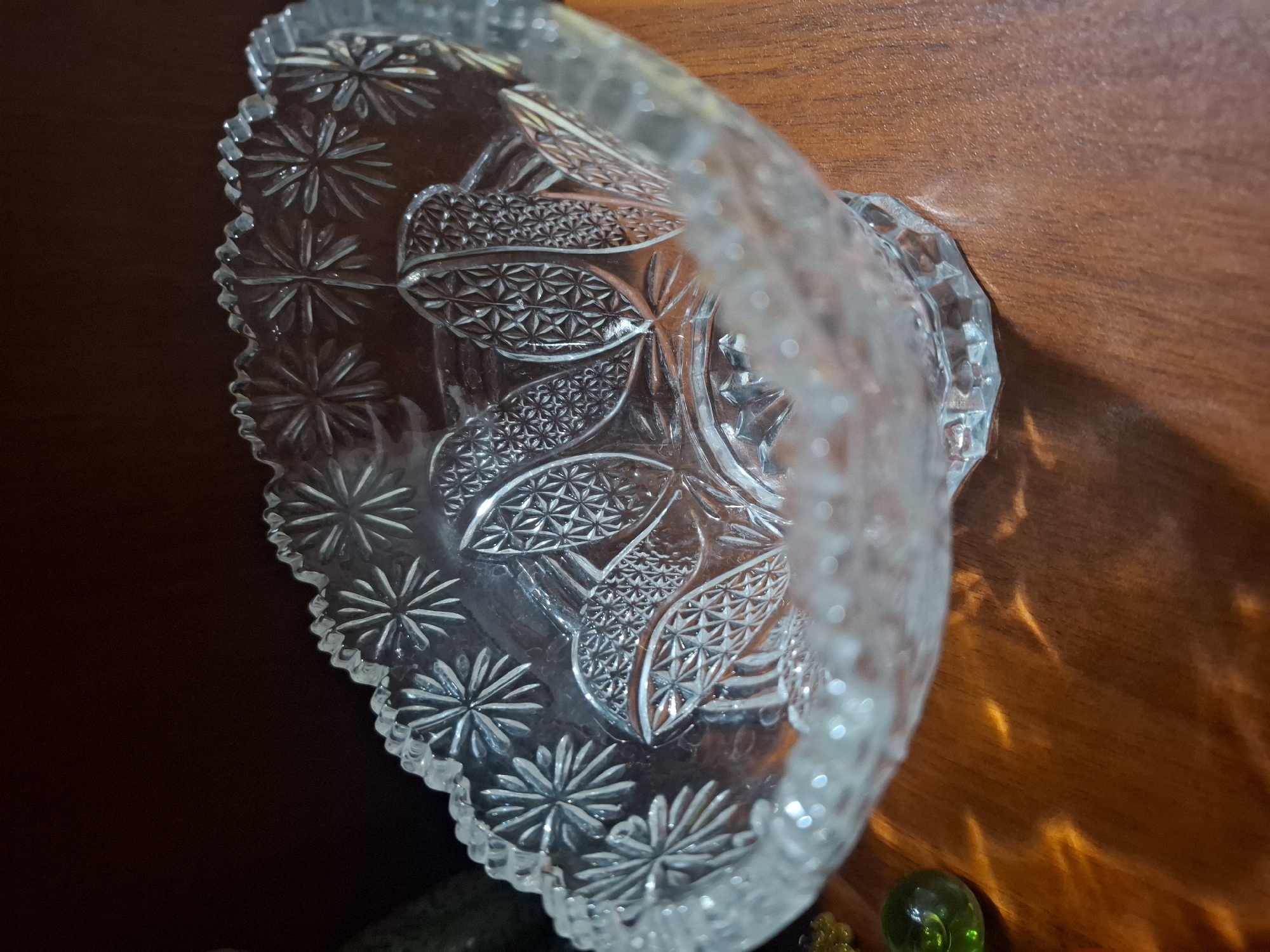 The image shows a close-up, eye-level view of a clear, ornate glass bowl with a fluted rim, resting on a polished wooden surface. The bowl is intricately decorated with starburst patterns and leaf-like motifs that have a textured, geometric design. Sunlight or lamplight is refracting through the glass, creating warm, abstract patterns of light and shadow on the wooden table. To the left of the bowl, a green glass marble is visible. The background is dark and out of focus, suggesting an indoor setting. The location is not discernible from the image itself.
