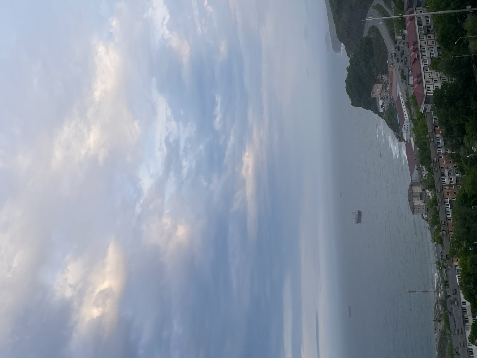 The image captures a broad coastal panorama, likely a bay or harbor in Vladivostok, Russia, under an overcast sky. The upper portion of the scene is dominated by a vast expanse of sky, filled with layered clouds ranging from soft white and light grey to darker, bluish-grey tones. Patches of brighter clouds suggest diffused sunlight filtering through.
The middle ground features a wide body of water, appearing grey-green with visible surface waves, indicating some movement or wind. Two distinct vessels are present: a larger, squarish ship or barge floats closer to the foreground, and a smaller, elongated ship is visible further in the distance.
The lower part of the image reveals a rugged, mountainous coastline adorned with a bustling town. Numerous multi-story buildings, many featuring red or dark-colored roofs, are clustered along the waterfront and extend up the green, tree-covered hillside. A winding road, with several vehicles visible, traces the contours of the land, connecting different parts of the settlement. Lush green trees and vegetation are interspersed among the urban structures and along the natural landscape.
The weather appears cloudy but not stormy, with daylight present, suggesting an overcast morning or afternoon. No people or visible text are discernible in the image.