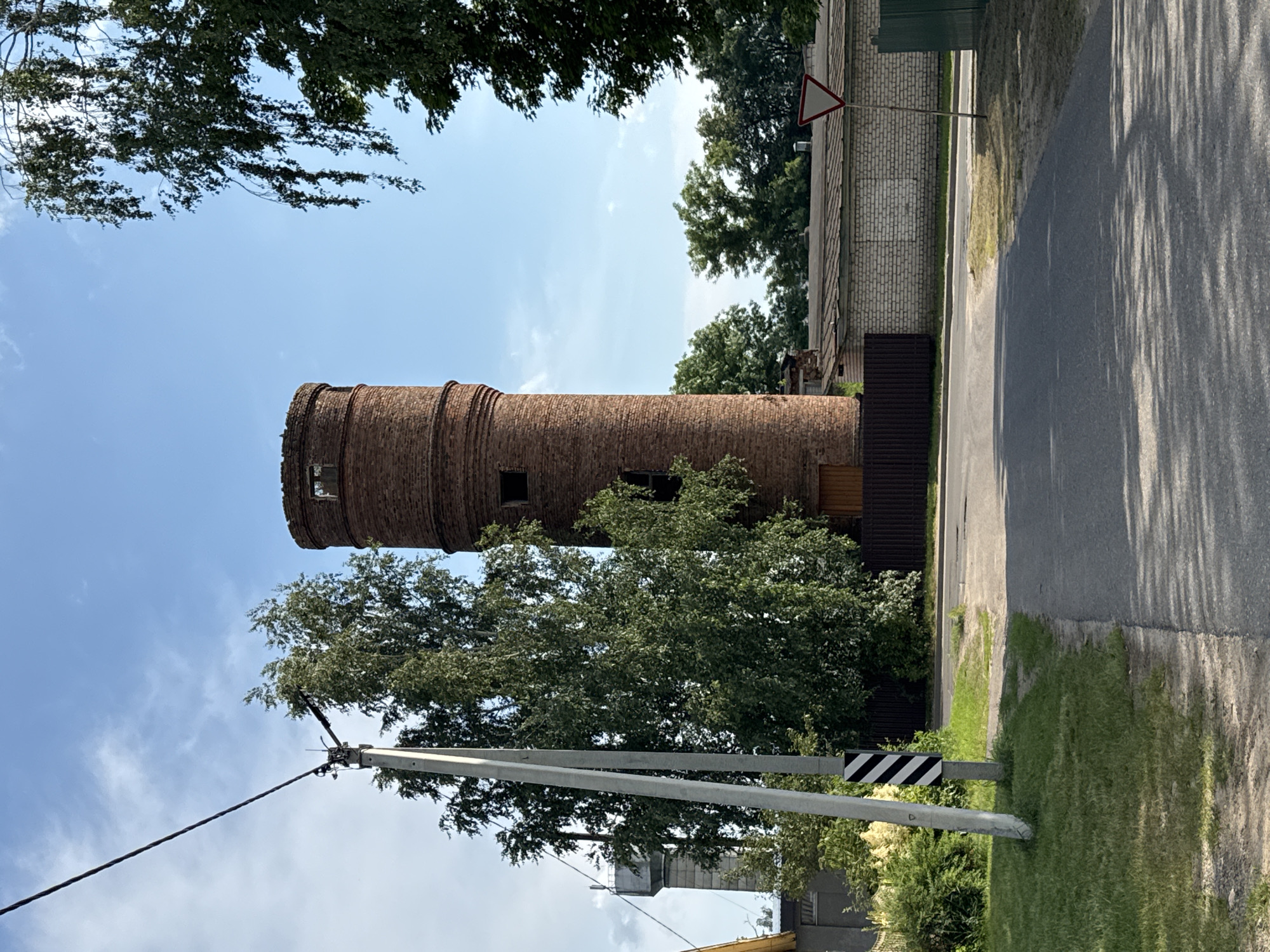 The image shows a red brick tower, possibly a water tower, with arched windows. The tower is surrounded by trees and greenery, with a road and a fence visible in the background. The sky is clear and blue, suggesting a sunny day. The overall scene appears to be in a rural or semi-rural area, possibly a village or small town. There are no people visible in the image. The photo is taken from a low angle, looking upwards towards the tower. The lighting and shadows suggest the time of day is mid-morning or early afternoon. The location is identified as Cherven, Belarus. There is a triangular warning sign visible on a pole, but no visible text.
