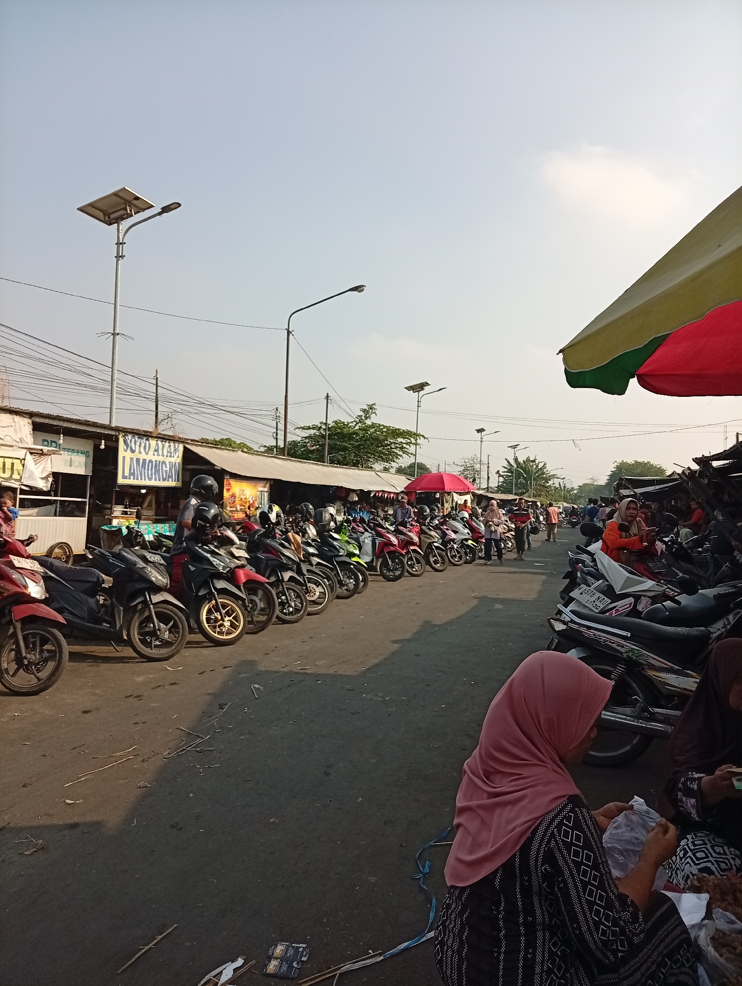 A low-angle, eye-level medium shot captures an outdoor marketplace in Larangan, Indonesia. The scene is dominated by a long row of parked motorcycles on a paved road. A few people are visible, including a woman in a pink hijab sitting on the ground to the right, holding plastic bags. Another woman in a dark hijab is seated next to her. Behind the motorcycles, stalls with awnings line the street, some with signs. One sign, in Indonesian, reads "SOTO AYAM LAMONGAN." Overhead, power lines crisscross the sky, and streetlights, including one with a solar panel, are mounted on poles. The sky is pale blue and clear. The ground is a mix of pavement and scattered debris, including dried leaves and twigs. The overall impression is of a bustling, everyday market scene under a bright sky.