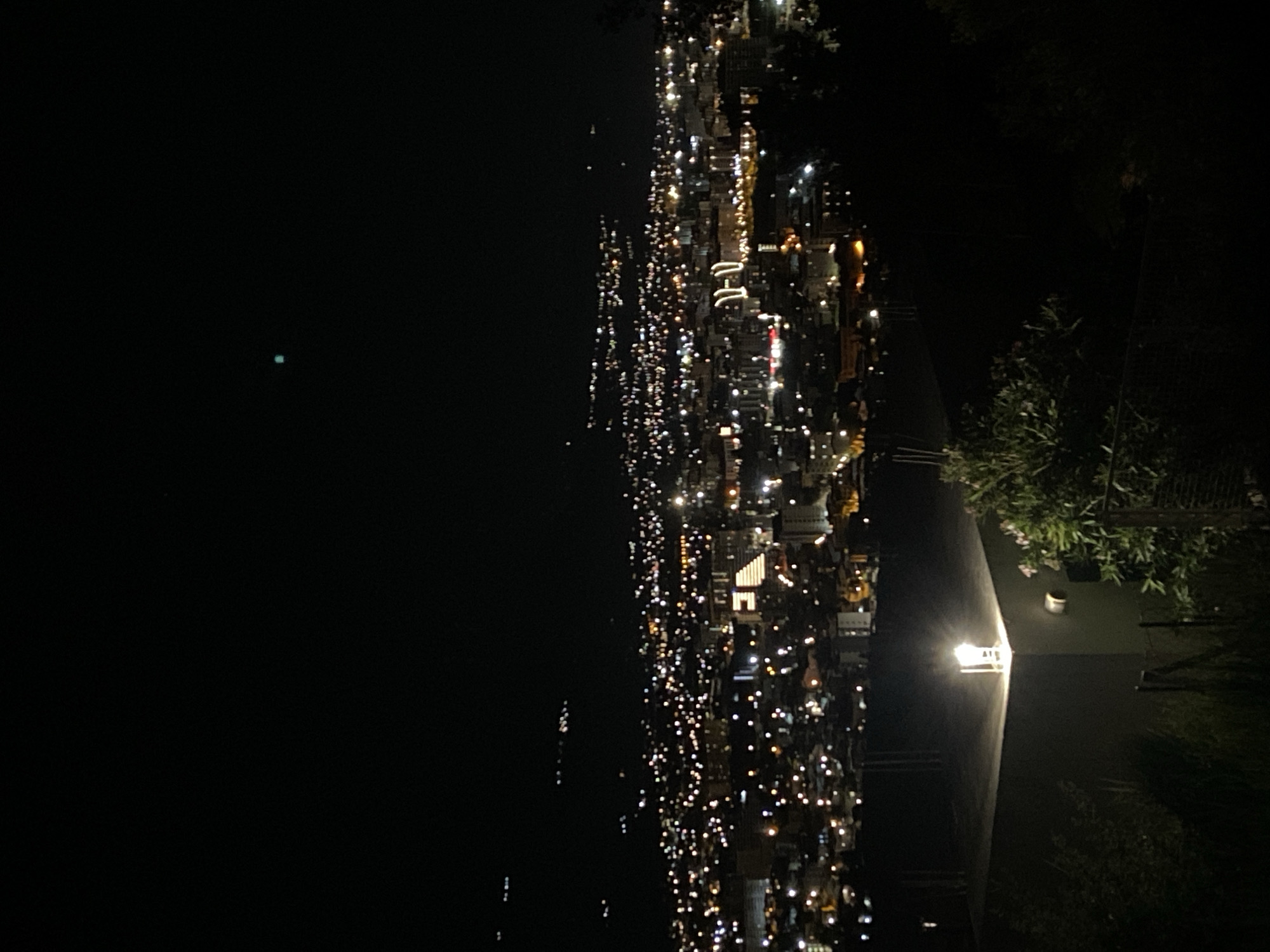 The image shows a nighttime cityscape, possibly in Šušanj, Montenegro, viewed from an elevated position. To the left, a dark expanse of water stretches out with a few faint lights visible in the distance. To the right, a densely packed urban area sprawls across the landscape, illuminated by numerous lights from buildings, streetlights, and possibly vehicles. The lights suggest a mix of building heights and types. On the foreground a lit lamp is visible next to plants.