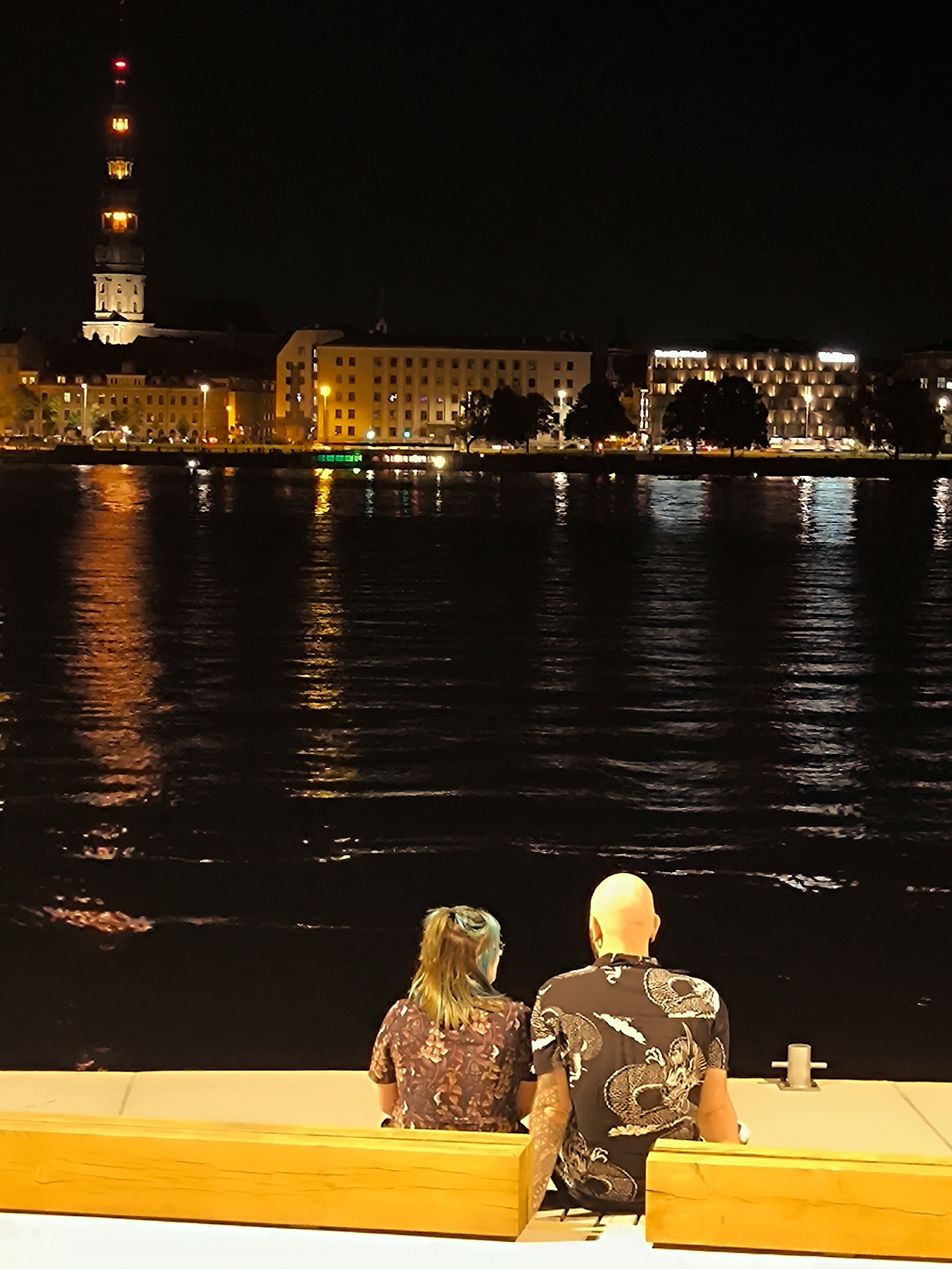 Two individuals, a woman with blonde hair and a man with a bald head, are seated on a bench at night, facing a body of water. The woman is wearing a patterned shirt. The man is wearing a black shirt with a white dragon design. In the background, across the water, is a city skyline illuminated by lights. A tall tower with lights is visible on the left side of the skyline. The water reflects the city lights. The scene appears to be a public waterfront area. The architecture of the buildings and the distinctive tower suggest the location is Riga, Latvia, likely the Daugava Riverfront with the St. Peter's Church spire in the background.