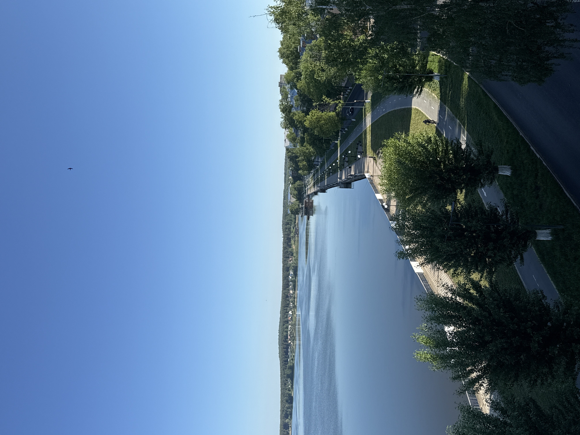 The image presents a high-angle view of a waterway, likely a river or large lake, in Votkinsk, Russia. Clear, blue sky occupies a significant portion of the upper-left of the image. Along the right side of the waterway runs a road and a pedestrian/bicycle path separated by a green strip. Trees line the edge of the path and road, suggesting an urban park setting. A cyclist is visible on the bicycle path. A bridge or similar structure crosses the waterway in the middle of the image. The opposite bank of the waterway shows signs of residential or urban development, with trees lining the shore. A small object, possibly a bird, is seen in the sky.