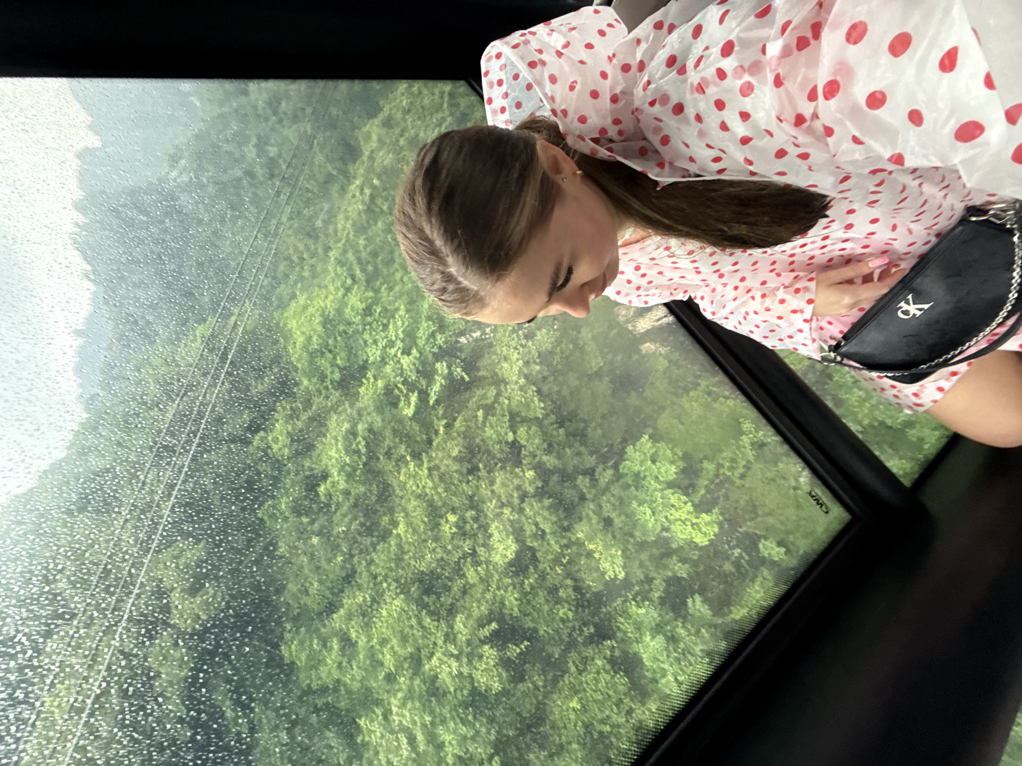 A woman is inside a cable car, possibly at Tianmen Mountain in Yongding District, China. Rain is visible on the cable car window, obscuring the view of the green, forested landscape outside. The woman has brown hair pulled back in a ponytail, and is wearing a white and red polka dot poncho. She has a small black purse with a silver chain.