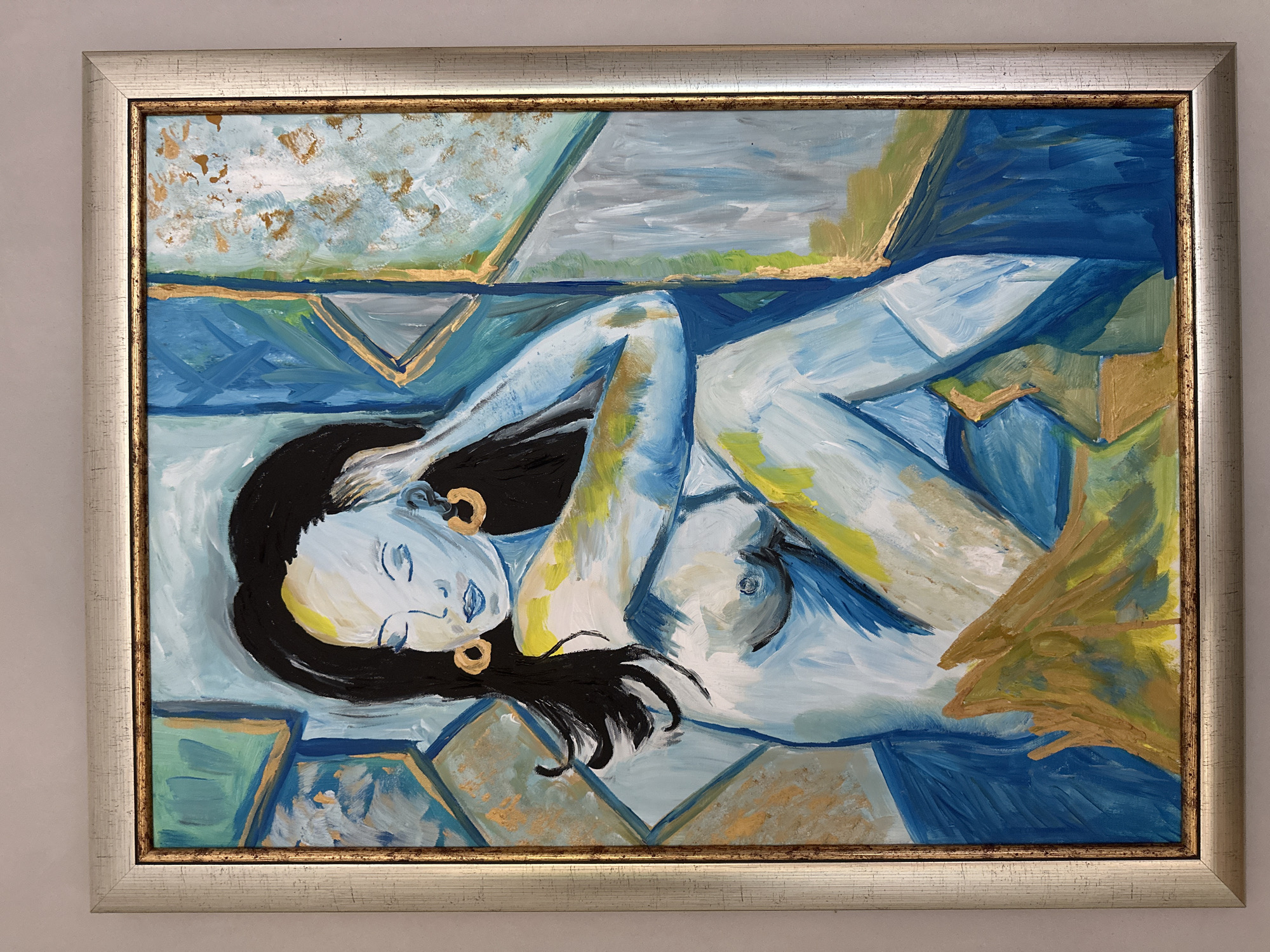 A framed oil painting depicts a stylized, nude figure of a person with dark hair and closed eyes, reclining with an arm raised to their head. The figure is rendered in shades of blue, white, and yellow, with touches of gold accents. They wear large, circular gold earrings. The background is an abstract arrangement of geometric shapes in blue, green, and gold. The painting is housed in a silver and gold ornate frame. The artwork is displayed against a plain, light-colored wall. The location is not discernible from the image.