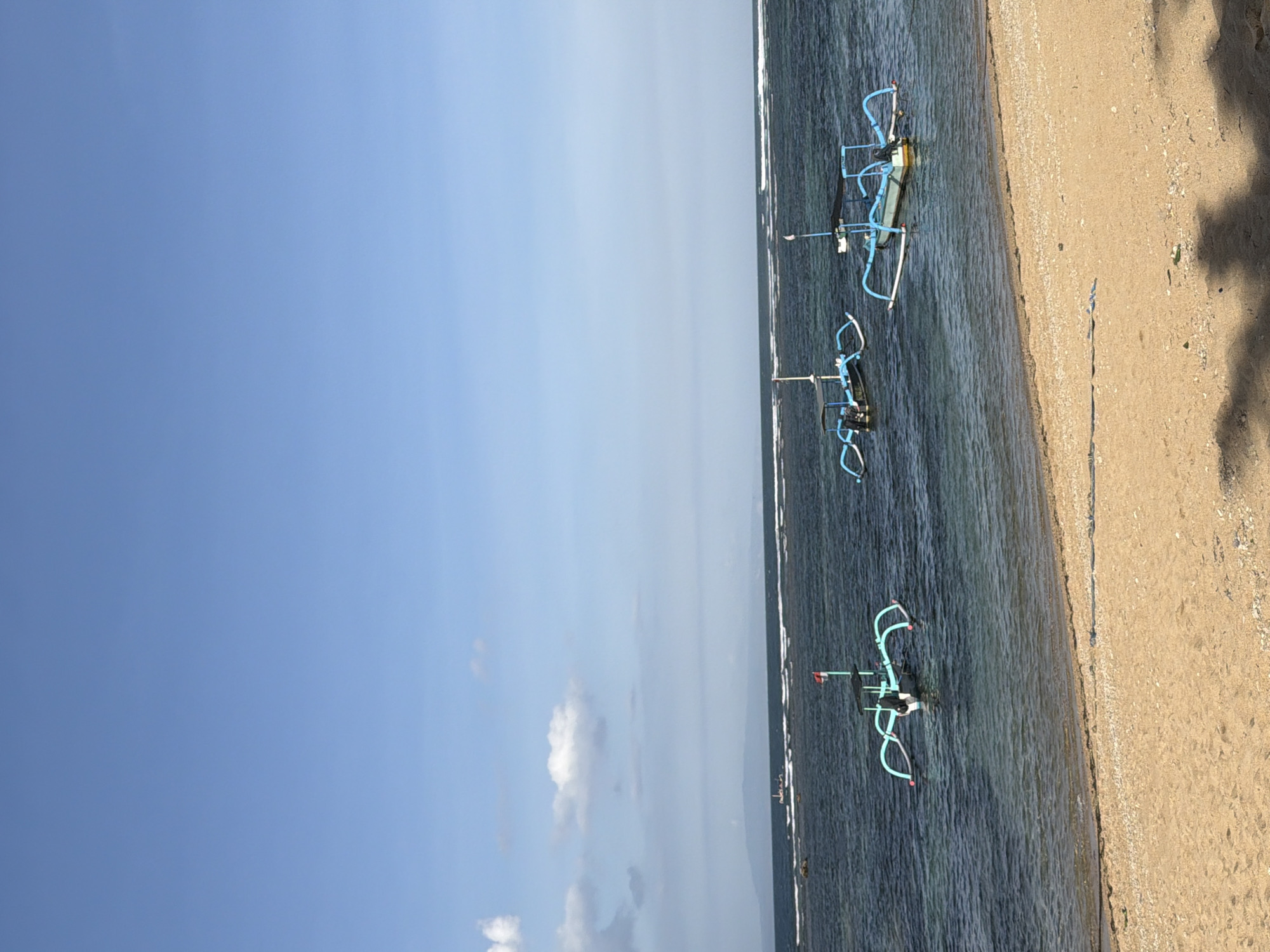 This media file shows a serene beach scene in Denpasar, Indonesia. The image is oriented vertically, with the sky occupying the left two-thirds of the frame and the ocean and beach on the right. The sky is a clear, pale blue with a few wispy clouds, suggesting a bright and possibly sunny day. The ocean is a deep, calm blue with gentle waves lapping at the shore.
Three traditional Indonesian jukung boats are visible in the water, near the sandy beach. These boats are characterized by their outriggers, which are painted a vibrant blue in the image. One boat is closer to the shore and appears to be grounded on the sand. The other two boats are further out in the water, floating gently. The sand on the beach is light brown and appears to be a mixture of fine grains and small pebbles, with some shadows cast upon it, possibly from palm trees or other vegetation not fully visible in the frame.
The scene evokes a sense of tranquility and natural beauty, typical of a tropical beach. The time of day appears to be either morning or late afternoon, given the quality of the light and the shadows. There is no visible text in the image.
