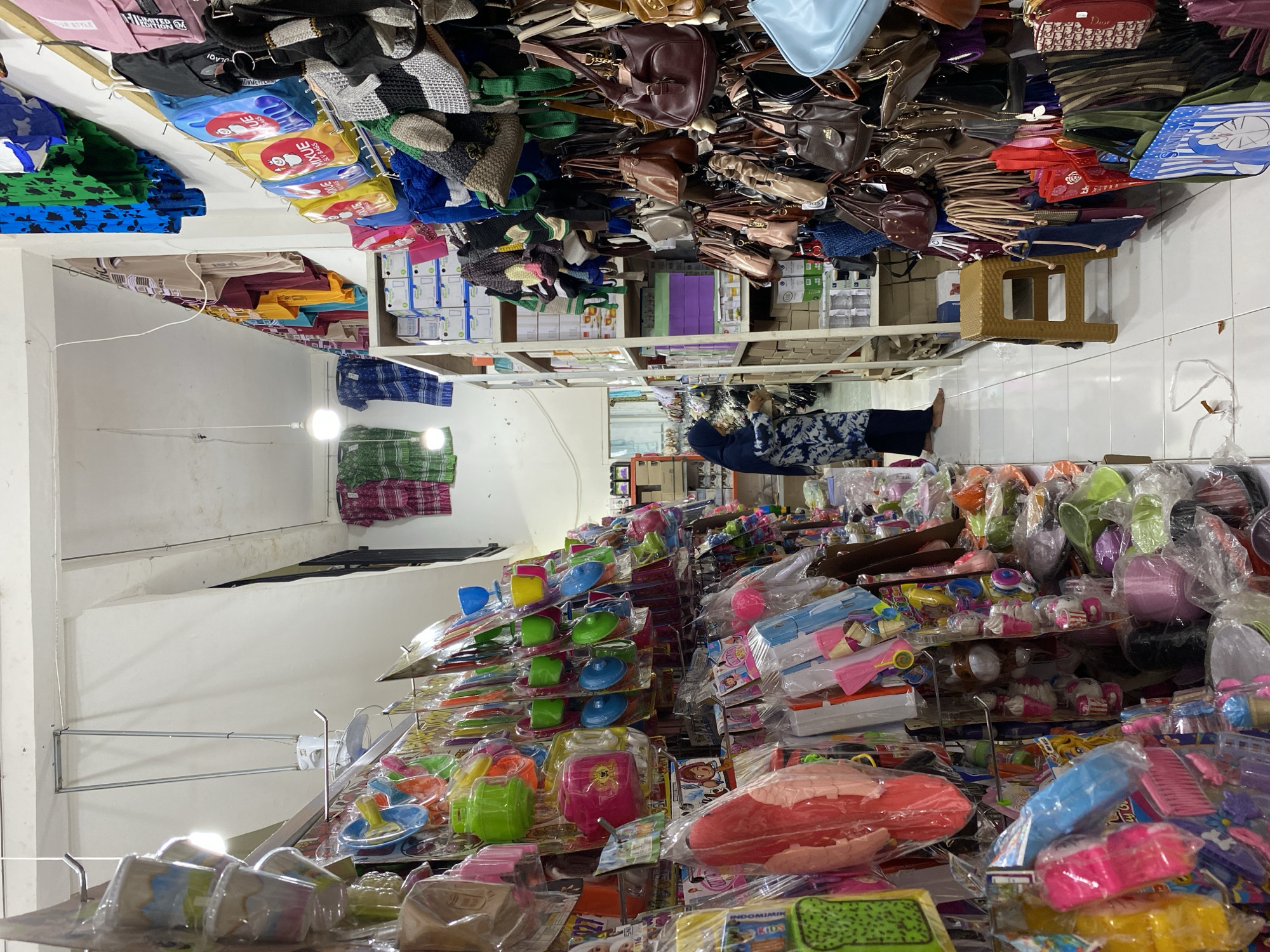 The image depicts the interior of a retail store in Lamteumen Barat, Indonesia, densely stocked with various merchandise. The store appears to have aisles lined with shelves and hanging displays. On the left side, racks are filled with packaged toys and kitchenware, including colorful plastic dishes and kitchen accessories. Further back, more shelves hold various items, and a woman wearing a hijab is seen examining products. The right side of the store features an extensive display of handbags and backpacks hanging from rods. These include a variety of styles, colors, and materials, such as leather, woven fabrics, and canvas. Shelving units behind the handbags contain boxes and additional merchandise. The lighting suggests an indoor environment, with overhead lights and a hanging fan visible. The floor is tiled.