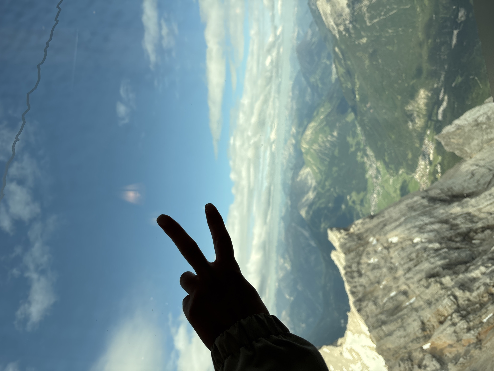 An extreme close-up shot captures a silhouette of a hand making a peace sign against a vibrant blue sky dotted with fluffy white clouds. The hand, positioned in the lower left corner, is a dark shadow with its index and middle fingers extended upwards, forming the iconic "V" shape. The background transitions from the sky to a panoramic view of rugged, green mountains and a valley speckled with buildings, suggesting a scenic mountainous landscape. The image has a slight tilt, adding a dynamic feel to the composition. There are some minor smudges or reflections on the surface through which the photo was taken, particularly in the upper right quadrant of the sky. The overall impression is one of a person enjoying a breathtaking view from a high vantage point, possibly in Ehrwald, Austria, given the landscape's characteristics.