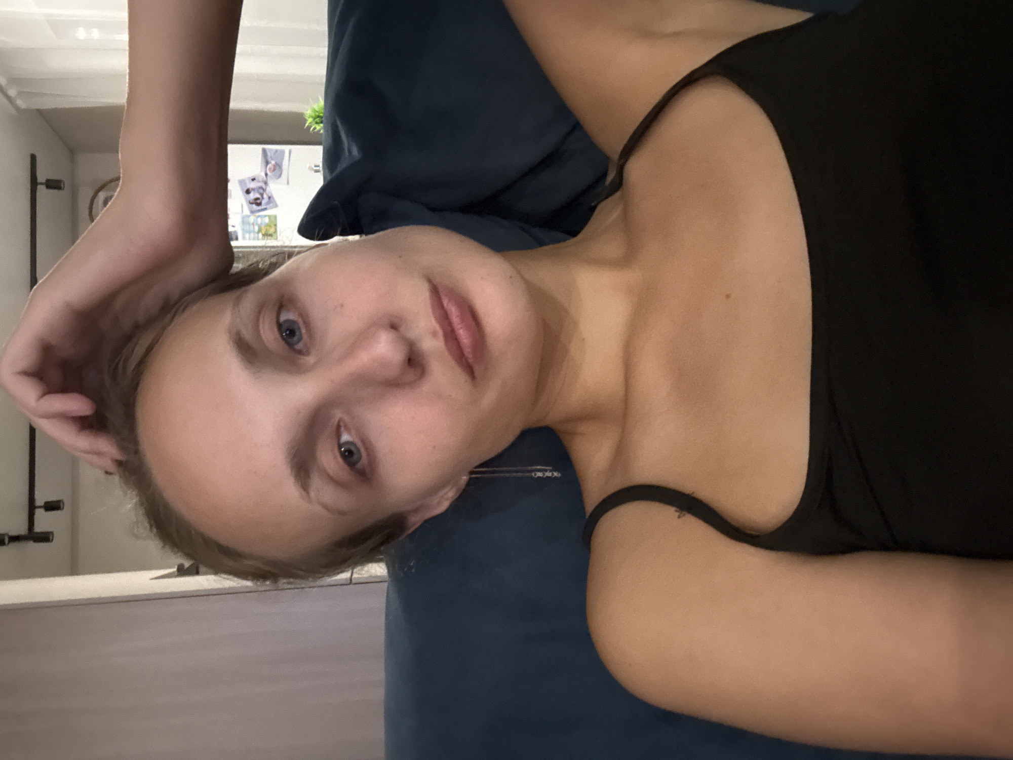 A young woman with fair skin and blonde hair is seated on a dark blue sofa, looking directly at the camera. Her left arm is raised, and her hand rests on the back of her head. She is wearing a black tank top. A delicate silver earring with small charms hangs from her left ear. In the background, a white appliance, possibly a refrigerator, is visible with several pictures attached to its surface. A green plant sits to the right of the appliance. The overall environment suggests an indoor setting, likely a living room. The location of Подшивалово, Russia, is not discernible from the visual elements present in the image.
