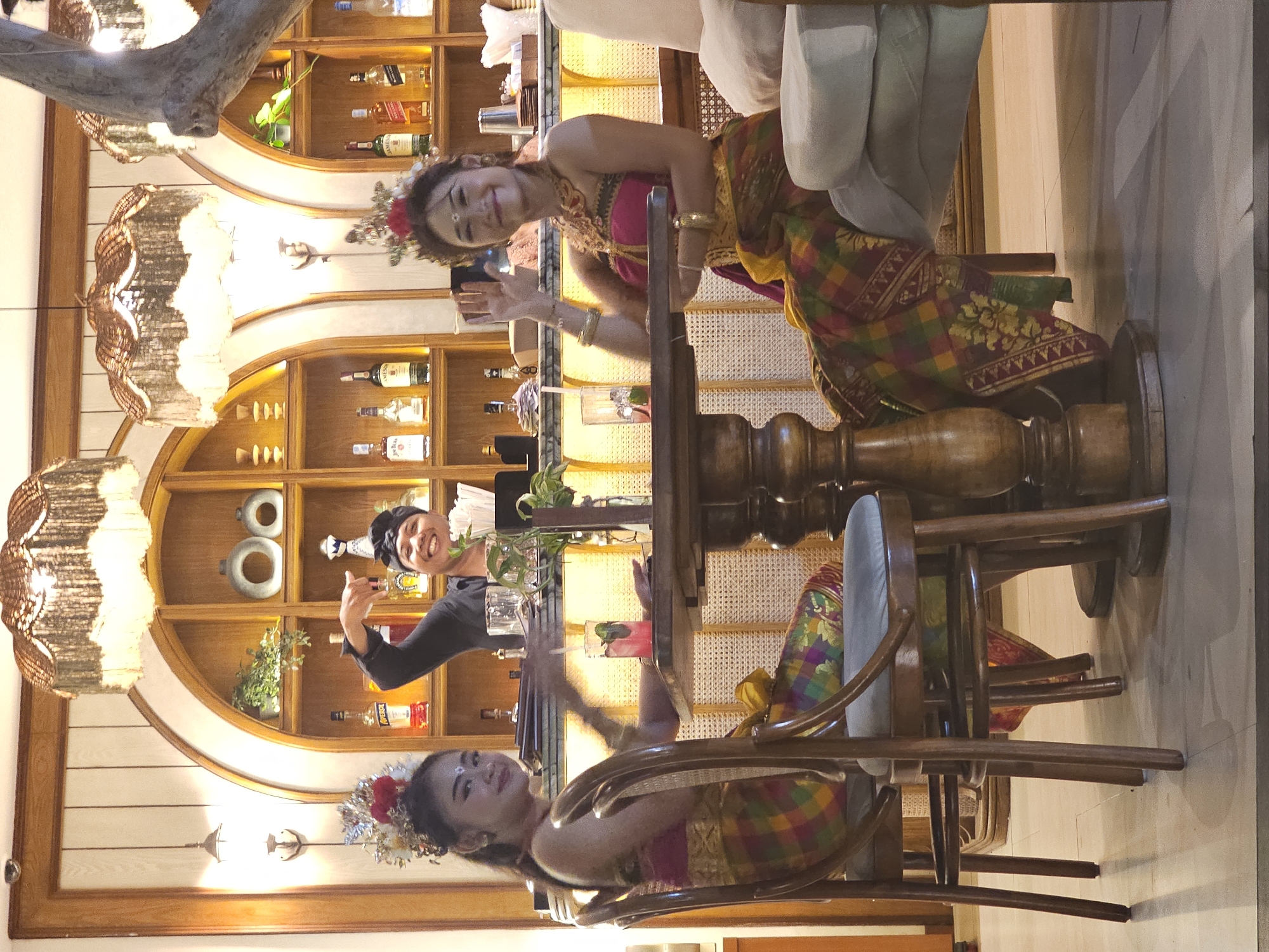 The image shows two women posing for a photograph at a table in a bar setting. The woman closest to the camera is wearing traditional Balinese clothing and a floral headpiece. She is smiling at the camera and raising her hand. Across the table, another woman is wearing a black top and a bandana. She is holding a cocktail glass, smiling, and giving a thumbs-up. The table has a glass of a pink drink with mint sprigs. A wooden bar and a shelf stocked with bottles of alcohol are in the background. The light fixture hanging from the ceiling has a woven design. The scene is well-lit and appears to be inside a bar or restaurant.
The location is Denpasar, Indonesia.