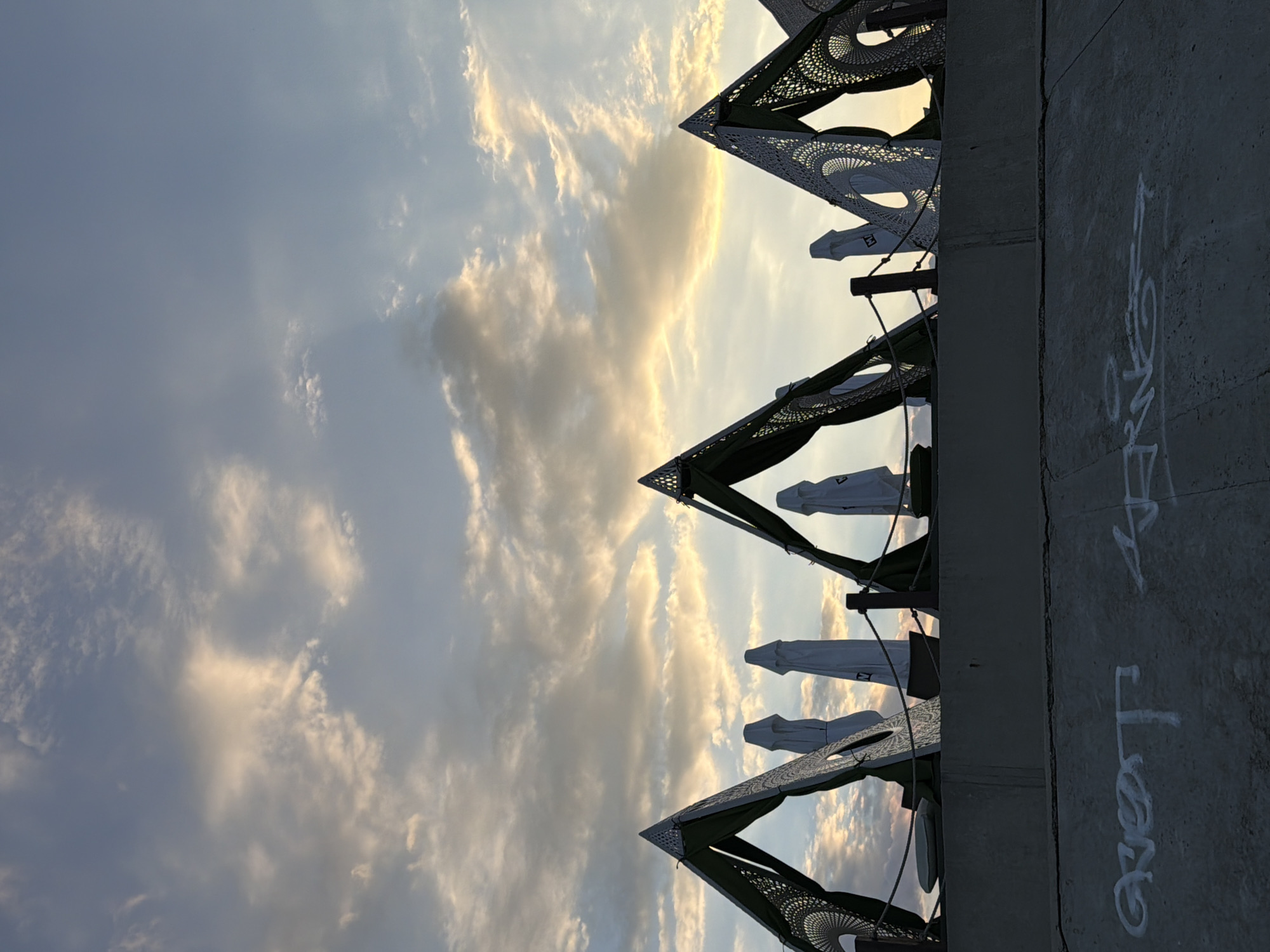 The image captures a sunset scene with a sky filled with scattered clouds, illuminated by warm golden light. In the foreground, there is a concrete structure with graffiti on its surface. Above this, a row of decorative, triangular canopies with intricate patterns is visible, each covered with a green fabric. Behind these canopies, several white, folded umbrellas are lined up. The scene is set in Fontanka, Ukraine, near a body of water, as suggested by the presence of beach-like umbrellas and canopies. The overall atmosphere is peaceful and serene, characteristic of a seaside resort at dusk.