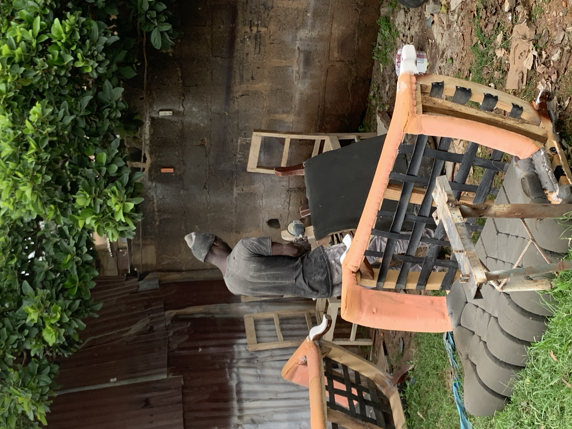 In Jos, Nigeria, a man wearing a grey beanie and a dark t-shirt is sanding a chair in an outdoor workshop. The scene is cluttered with furniture parts, including an orange-upholstered chair frame in the foreground and a disassembled seating unit. A corrugated metal structure is visible in the background, along with a brick wall and lush green foliage. The ground is covered in grass, debris, and some construction materials.