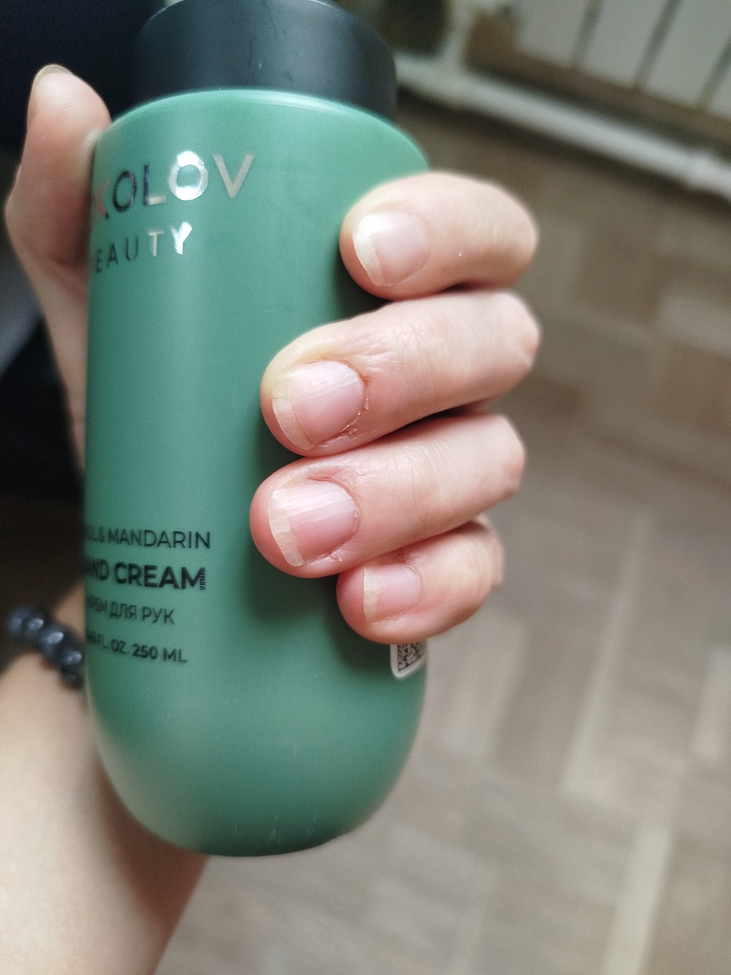 In this close-up, medium shot, a person's hand with short, unpolished fingernails gently holds a teal-colored bottle of hand cream. The bottle features silver lettering, partially obscured by the hand, that reads "KOLOVO BEAUTY" and "& MANDARIN HAND CREAM". Below this, in smaller text, are the Cyrillic words "КРЕМ ДЛЯ РУК" and the volume measurement "6.7 FL. OZ. 250 ML". A beaded bracelet is visible on the wrist of the person holding the bottle. The background is blurred, showing a light-colored, textured surface that suggests an indoor setting. The overall lighting is soft and natural. This image was taken in Zavodoukovsk, Russia.