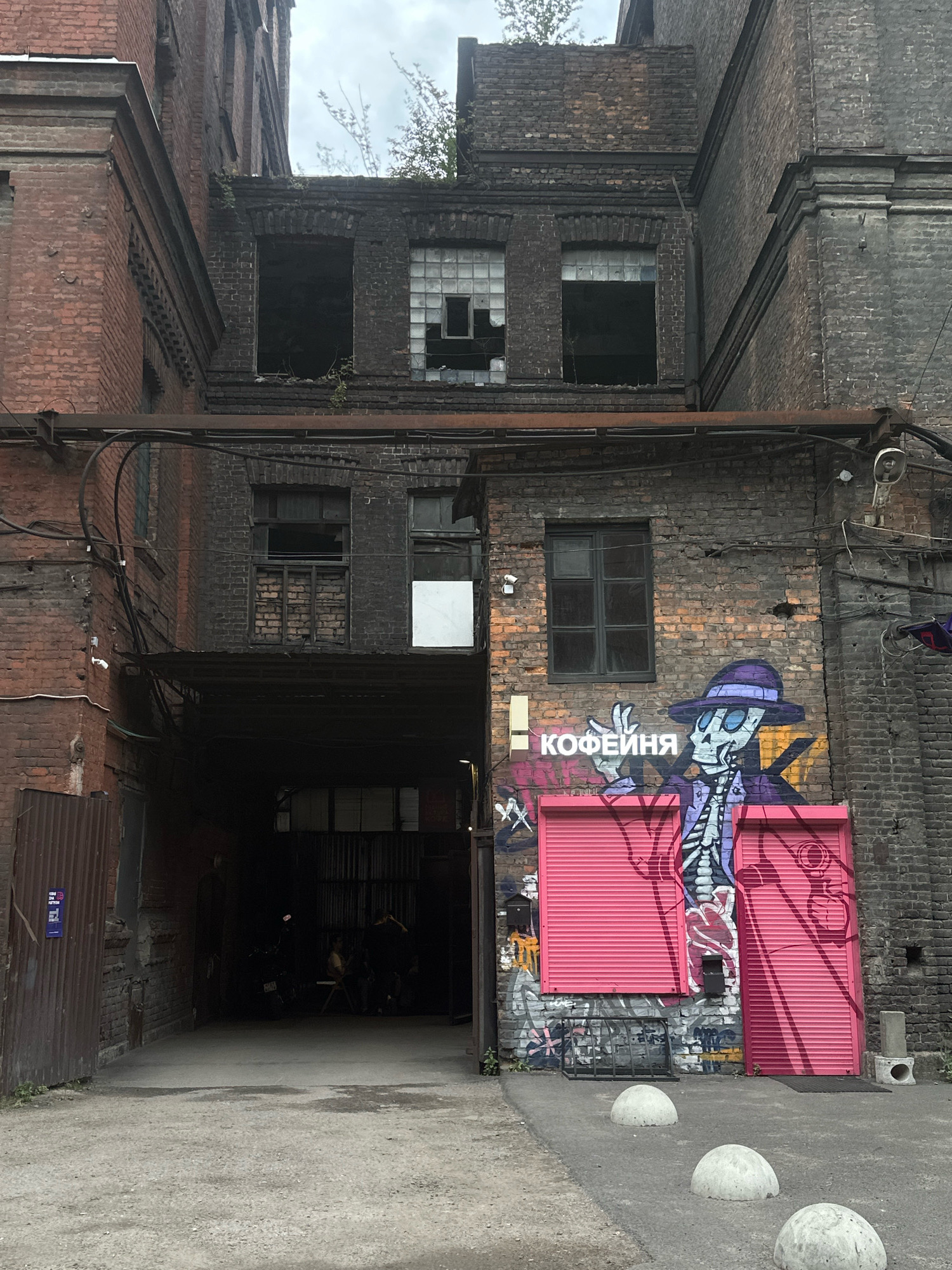 This image depicts a gritty, urban courtyard in Saint Petersburg, Russia, likely an industrial or former industrial area, given the weathered brick buildings and utilitarian architecture. The courtyard itself is paved with gravel and asphalt, with several concrete bollards lining the foreground.
Dominating the right side of the frame is a brick building with a dark, grimy facade. A vibrant graffiti mural of a skeleton in a purple hat and trench coat, holding a coffee cup, covers a section of the wall. The text "КОФЕЙНЯ" (meaning "coffee house") is written in neon pink above the skeleton. The mural is painted over closed pink roller shutters. Below the shutters, there are more graffiti tags and a metal grate.
To the left of the mural, a large, dark archway leads into another part of the courtyard. Inside the archway, the lighting is dim, and silhouettes of people and what appear to be motorcycles can be seen. A corrugated metal fence is visible on the far left, adjacent to another brick building.
The buildings are multi-story structures with exposed pipes and wiring running along the facades. Many windows are either broken, boarded up, or covered with glass blocks, suggesting a state of disrepair or abandonment. Vegetation is growing on the ledges and rooftops of the upper floors, adding to the sense of decay and nature reclaiming the urban environment. The sky is overcast.