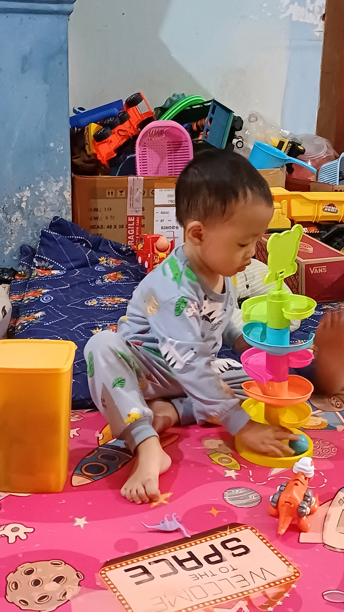 A young child is playing with a multi-colored toy spiral tower and a small orange dinosaur toy on a pink mat. The child, wearing light blue pajamas with a zebra pattern, is sitting on the floor, with their right hand reaching towards the toy spiral. The mat is decorated with images of planets, stars, and rockets, and the words "WELCOME TO THE SPACE". In the background, there are several other toys, including a red truck, a pink basket, and various boxes. The scene appears to be indoors, likely in a residential setting in Wonokerso, Indonesia.