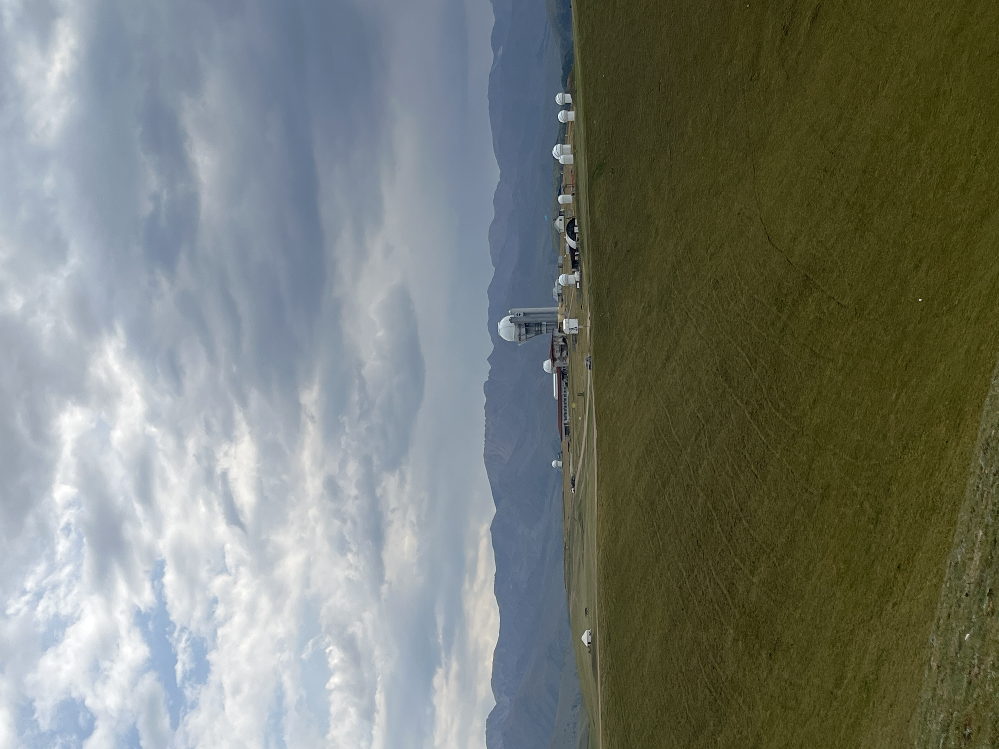 An aerial view captures a vast, undulating green grassland under a cloudy sky. In the mid-ground, a complex of buildings is situated on a plateau. This complex includes several dome-shaped structures, a tall, slender building topped with a dome, and a longer, red-roofed building. The area appears to be an observatory or research facility. Mountains form a distant, hazy backdrop to the scene. The environment is natural and rural, with no visible people or animals. The scene is static and devoid of action.