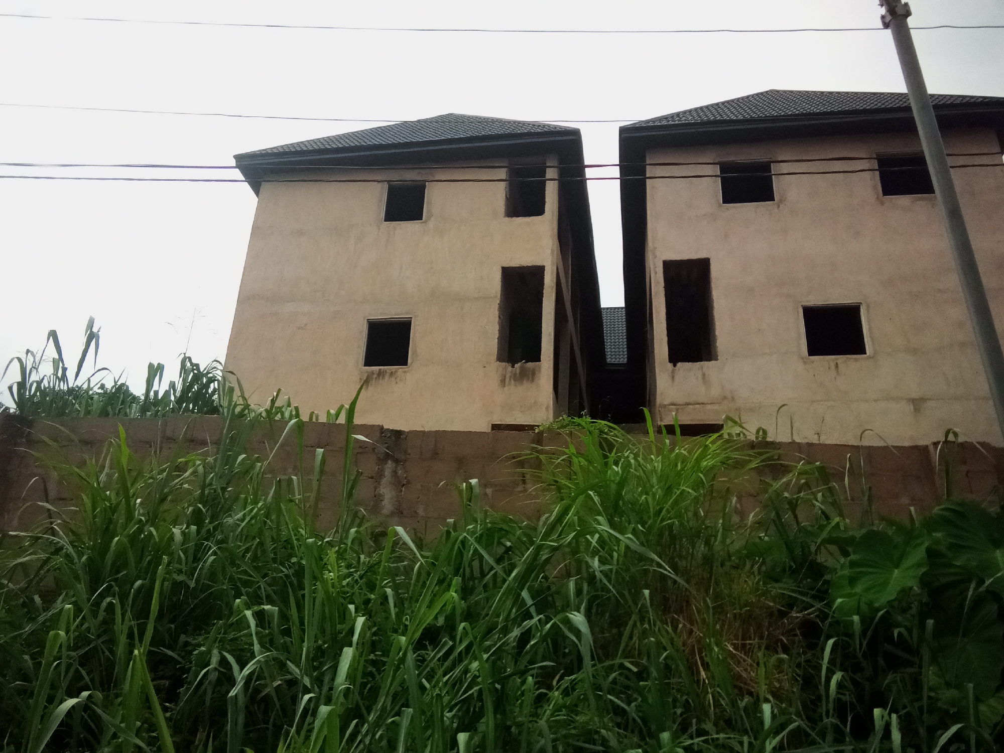 The image shows the exterior of two unfinished, multi-story buildings in Oko Town, Nigeria. The buildings are made of light-colored concrete and have dark, tiled roofs. Both structures have visible windows and doorways that are currently empty, indicating construction is ongoing or has been halted. The buildings are positioned closely together, with a narrow gap between them. In the foreground, dense green vegetation, including tall grasses and broad leaves, grows in front of a low, weathered concrete wall. Overhead, power lines stretch across the sky, and a utility pole is visible on the right side of the frame. The sky is overcast, suggesting a cloudy or hazy day.