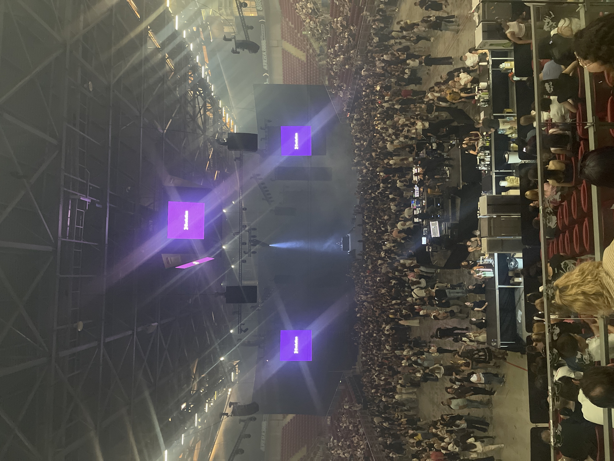The image depicts a large indoor event, possibly a concert or conference, at Arena Armeec in Sofia, Bulgaria. A substantial crowd is gathered in front of a stage with multiple screens displaying a purple logo of "3DataVideo". Lighting rigs and support structures are visible in the arena's ceiling. The attendees are densely packed on the floor level and in the tiered seating areas. The image is taken from a higher vantage point, likely from the seating area. The overall atmosphere seems lively and energetic.
