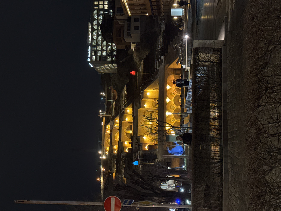 The image captures the Yerevan Cascade at night in Yerevan, Armenia, presenting a multi-tiered architectural complex illuminated against a dark sky. The Cascade ascends a hillside, featuring numerous steps, retaining walls, and a series of arched structures, all brightly lit with warm yellow-orange light that casts distinct shadows. Integrated into the complex are various sculptures, including a tall, slender blue human-like figure on the left side of a lower tier and a similar red human-like figure on a higher tier to the right. A prominent modern building with the "ServiceTitan" logo visible on its rooftop stands illuminated on the hilltop above the Cascade, to the right.
In the mid-ground, a paved pedestrian area is separated from the viewer by a low, dark hedge. A person in dark attire is partially visible behind this hedge, facing towards the illuminated Cascade. Further to the right, a second individual in a dark coat and hat stands near a light-colored sculpture of a reclining female figure. Additional, less distinct human figures are visible on various levels of the Cascade steps.
The immediate foreground features dark cobblestone paving and low, sparse shrubbery. On the far left, a tall utility pole supports a circular red "Do Not Enter" sign positioned above a blue directional arrow sign. Bare-branched trees are interspersed throughout the lower and mid-levels of the Cascade and surrounding areas. Streetlights and ambient building illumination contribute to the scene's lighting.