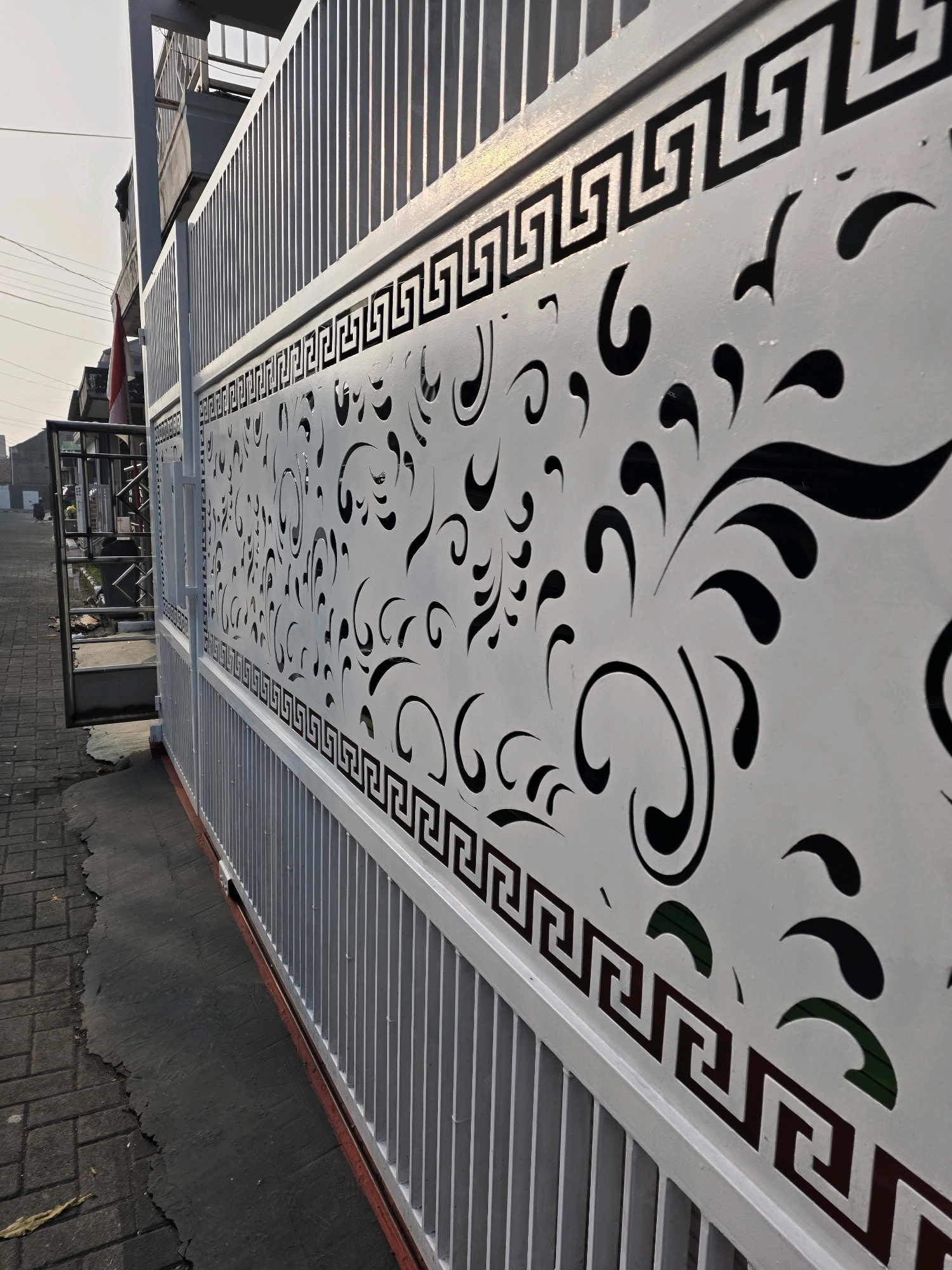 The image depicts a white metal gate with intricate laser-cut patterns in Bojongsoang, Indonesia. The gate features a Greek key border above a swirling, floral-inspired design, with negative space creating the patterns. Vertical metal bars form the upper and lower sections of the gate. To the left, a portion of an opened gate reveals a darker metal structure and a red and white Indonesian flag. The ground in front of the gate is paved with dark gray rectangular bricks, transitioning to asphalt with some damage. In the background, a building with multiple stories and windows is visible, along with power lines and a hazy sky. The lighting suggests it is daytime, possibly late afternoon due to the long shadows.