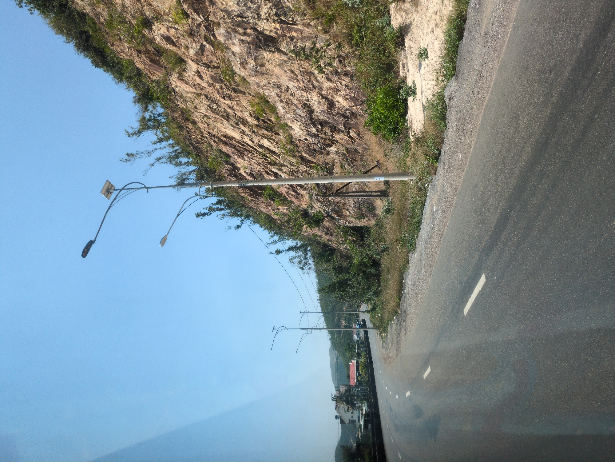 The image shows a section of asphalt road with white lane markings, alongside a rocky cliff face covered in vegetation. Streetlights line the road. In the distance, buildings and hills are visible. The scene appears to be in Hòa Mỹ, Vietnam.