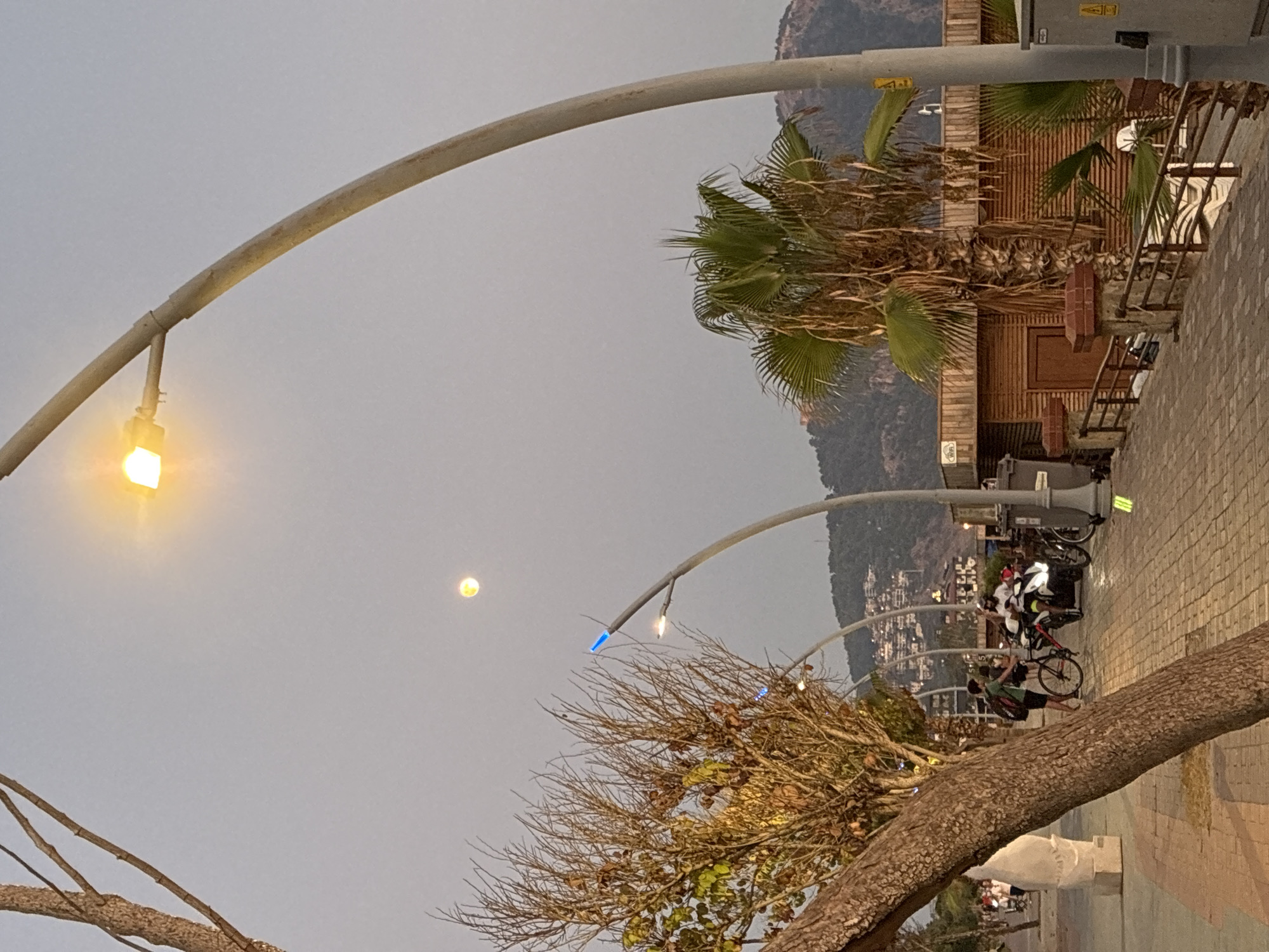 The image depicts a street scene in Alanya, Turkey, during dusk. A prominent curved lamppost with an illuminated light is positioned in the foreground. Further down the walkway, another lamppost with blue lights is visible. The sky is overcast, with a faint moon visible. The walkway is paved with interlocking bricks, and a tree with sparse foliage stands to the left. In the middle ground, two individuals are present; one is standing next to a bicycle, and the other is on a scooter. Beyond them, a low-rise wooden building with a sloped roof is situated. In the background, a hillside covered with buildings and vegetation rises. The overall atmosphere suggests a mild evening in a coastal city.