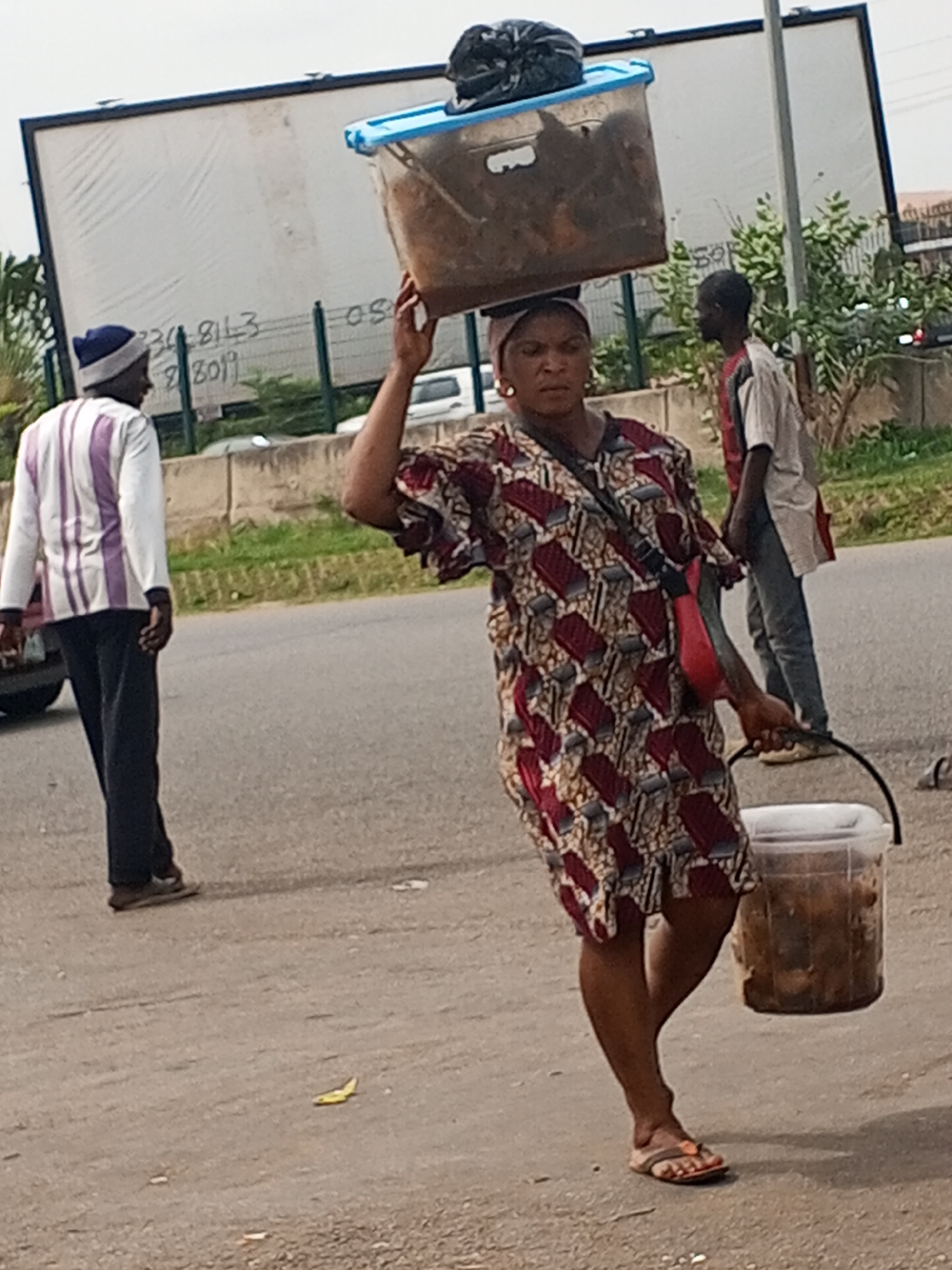 A woman is walking down a street in Abuja, Nigeria. She is carrying a large container on her head and a bucket in her hand. The container on her head is blue and clear, and it appears to contain food of some sort. The bucket in her hand is also clear and appears to contain food. She is wearing a brown, white, and red patterned dress, sandals, and earrings. She has a red purse slung across her shoulder. Two men are walking nearby. A building with a large white sign is visible in the background. There is also some greenery and a barrier behind the sign. The street appears to be a busy road. There is litter on the street, including a banana peel.