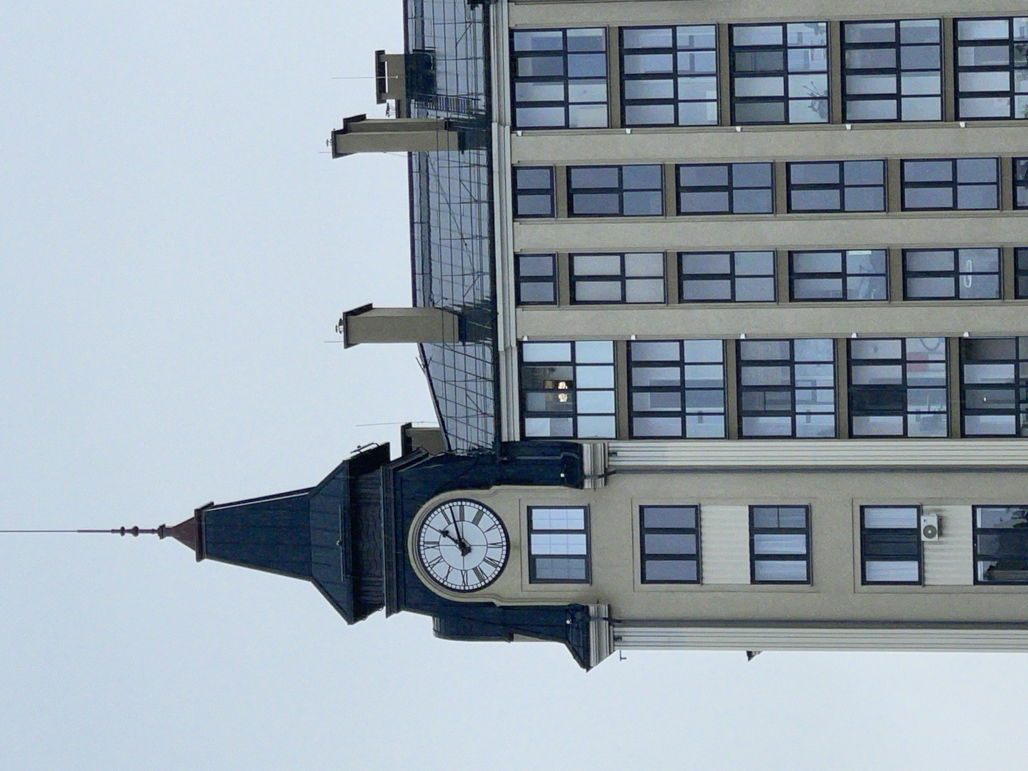 The image shows the facade of a modern apartment building in Zelenogradsk, Russia. The building features a prominent clock tower with a dark pitched roof and a spire. The clock face is round with Roman numerals and black hands. Below the clock are several stories of windows with dark frames. To the right of the clock tower, the building continues with a light-colored facade and a series of uniformly arranged windows with dark frames. The roof of this section is partially covered with glass. Two large chimneys are visible on the glassed roof. The sky in the background is overcast. The overall impression is of a contemporary residential building with classical architectural elements incorporated into its design.