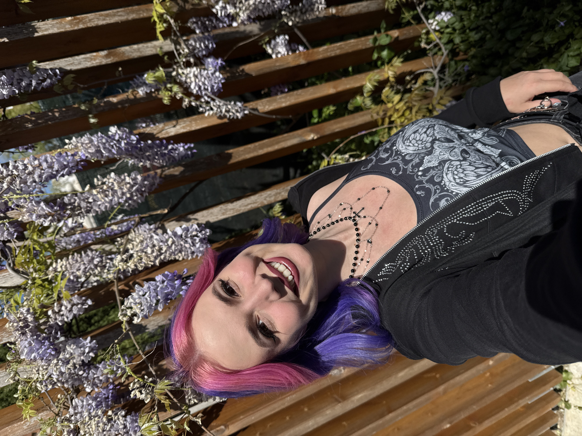 A fair-skinned young adult woman with pink and purple hair is seen in a slightly tilted medium shot. She is smiling and looking directly at the camera. She is wearing a black tank top with a grey paisley-like design, a black cross necklace, and a black jacket with silver embellishments. She is sitting on a wooden slatted structure, potentially a bench or trellis. Purple wisteria flowers are draped over the wooden slats. The scene appears to be outdoors in a garden or park setting. The location of the scene is not possible to deduce from the context of the image.