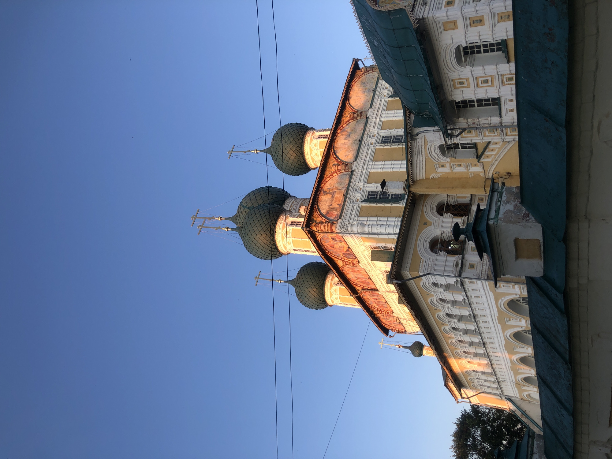 The image captures a low-angle view of the Assumption Cathedral in Tutaev, Russia, bathed in the warm, golden light of the setting sun. The cathedral's distinctive green domes, topped with crosses, are silhouetted against a clear, bright blue sky. The yellow and white facade of the building is adorned with intricate architectural details, including a row of arched windows and decorative white plasterwork. A dark green, corrugated metal roof partially obscures the lower section of the cathedral. In the foreground, a dark green, weathered fence runs horizontally across the bottom of the frame, with a portion of a white wall beneath it. Power lines are visible stretching across the upper portion of the sky, and a few scattered birds can be seen as tiny specks against the blue. The overall impression is one of serene grandeur, emphasizing the architectural beauty of the cathedral under the soft glow of twilight.