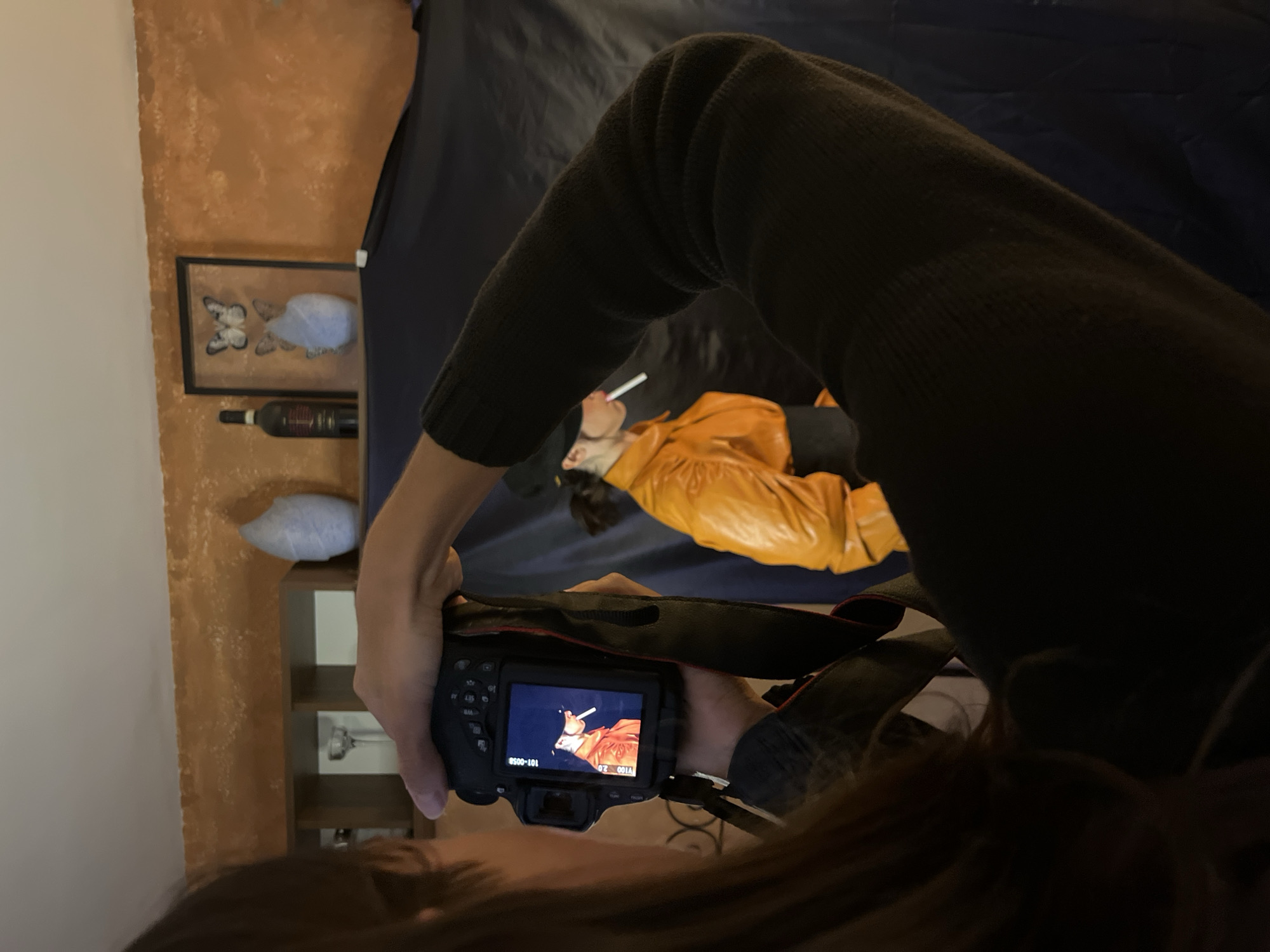 A person wearing a dark green sweater is holding a camera and taking a picture of another person lying on a dark blue surface wearing a yellow jacket and holding a cigarette. In the background, there is an orange-painted wall with a framed picture of butterflies, a wine bottle, a white pillow, and a wooden shelf. The location is not explicitly mentioned in the image and the city of Barcelona, Spain cannot be confirmed.