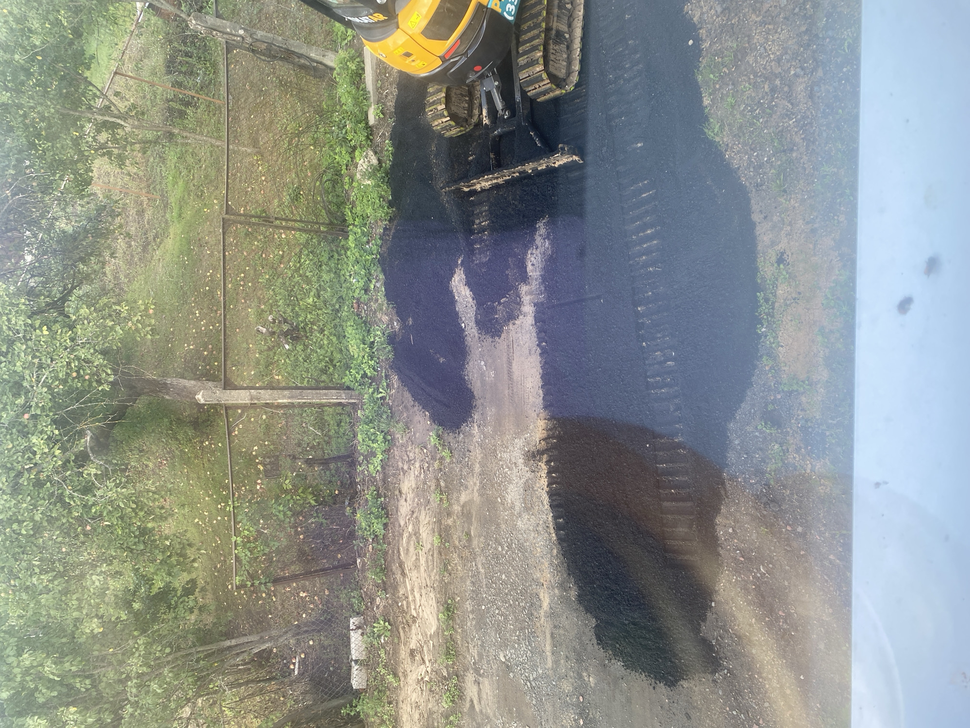 In Krupki, Belarus, an excavator with a yellow body and black tracks is spreading asphalt on a dirt and gravel surface. The excavator has a metal blade attachment at the front, which is in contact with the asphalt. A pile of loose asphalt is visible to the right of the excavator. The area appears to be outdoors, with vegetation and a fence in the background. Several apples are scattered on the ground under trees behind the fence. The foreground is partially obscured by a reflection or glare.