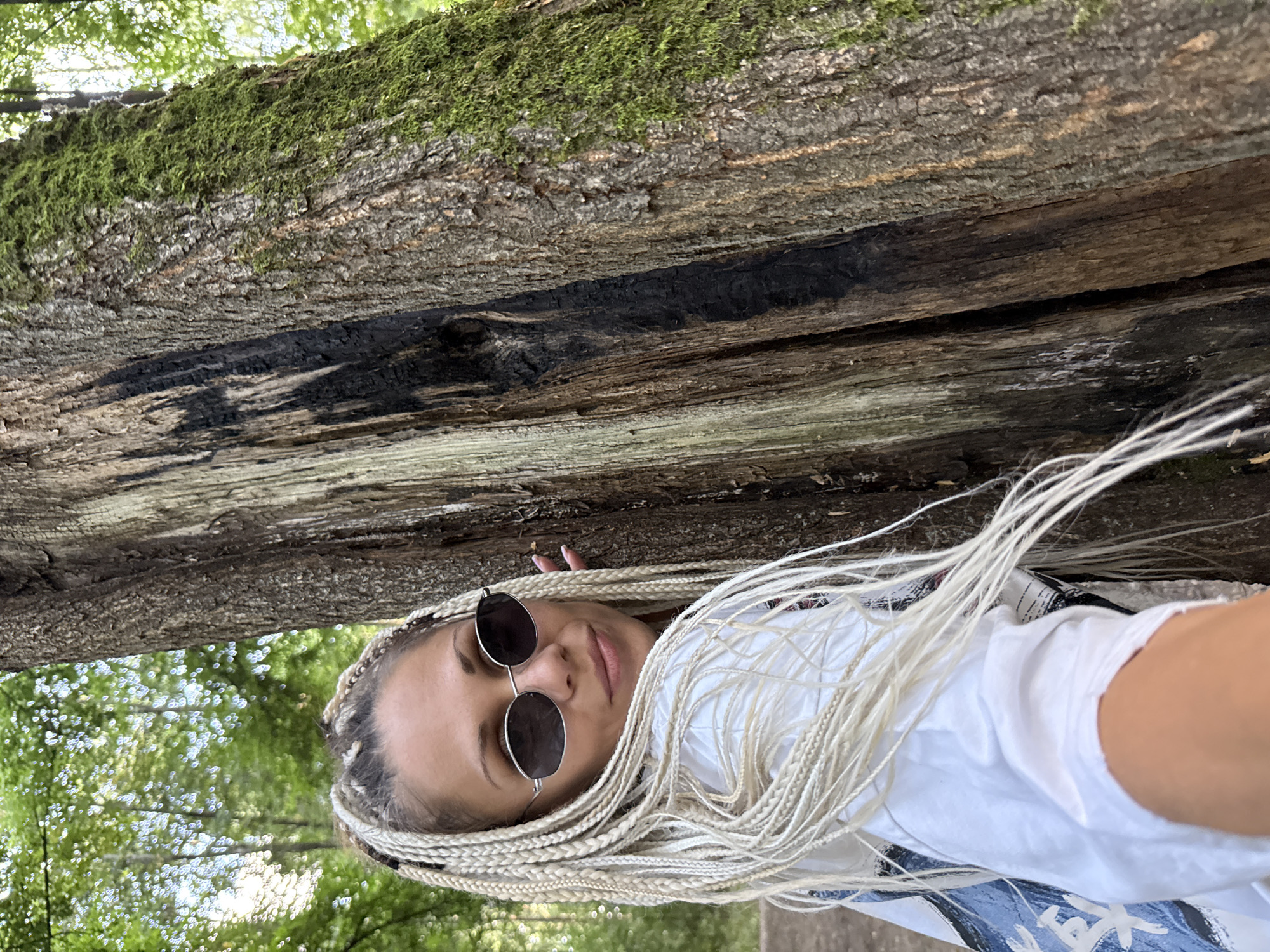 In Rusanovo, Russia, a woman with blonde braided hair and sunglasses poses next to a large, textured tree trunk. The tree exhibits a split down its center, with one side covered in moss and the other showing weathered, darker wood. The woman is wearing a white t-shirt and a backpack, her left hand partially visible in the foreground. The background reveals a lush green forest with dappled sunlight filtering through the leaves.