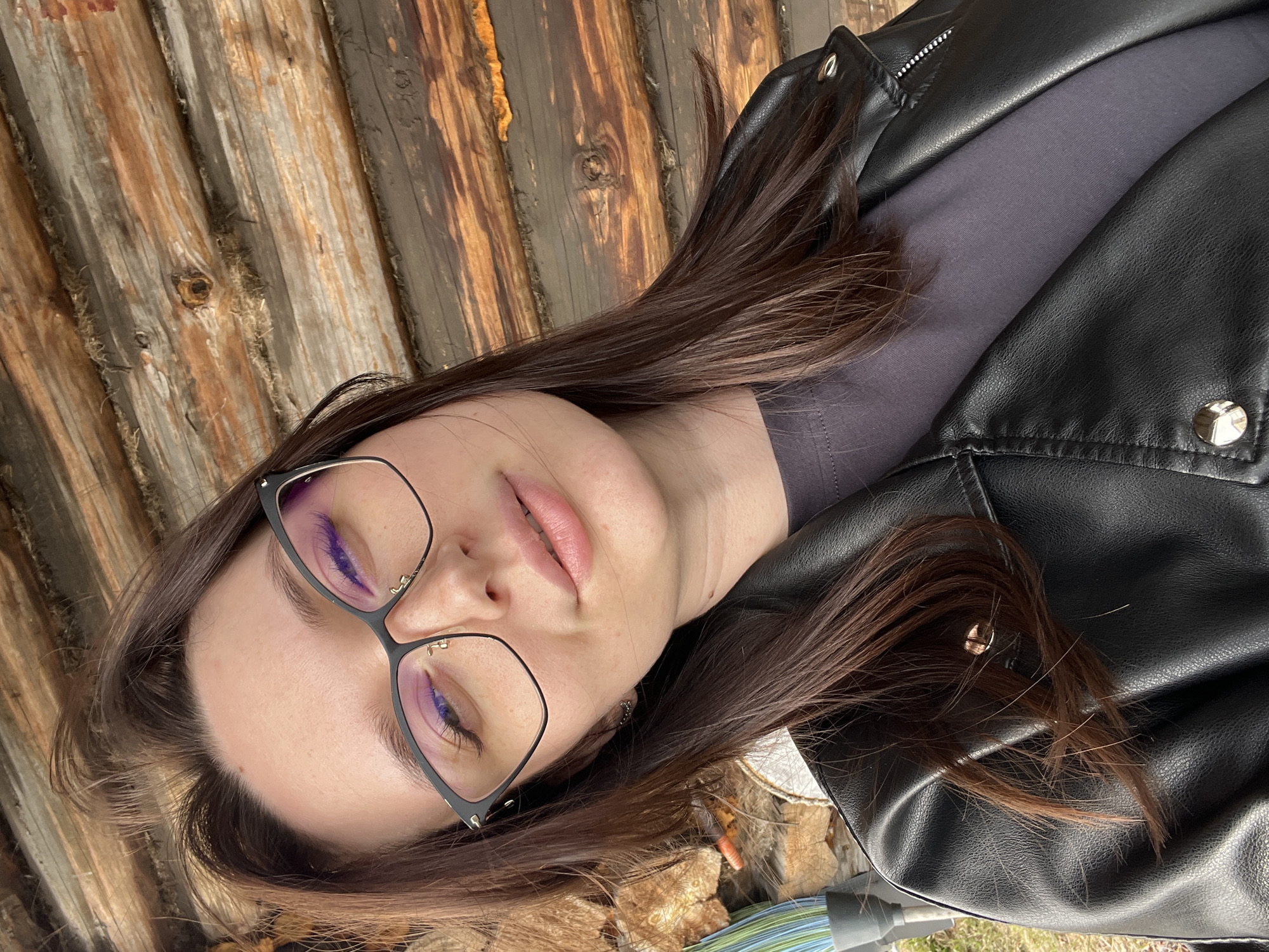 The image shows a woman wearing black-rimmed glasses and a black leather jacket. She is lying down, with her head tilted to the side. The background consists of rough wooden planks. The image location is Usol'tseva, Russia.