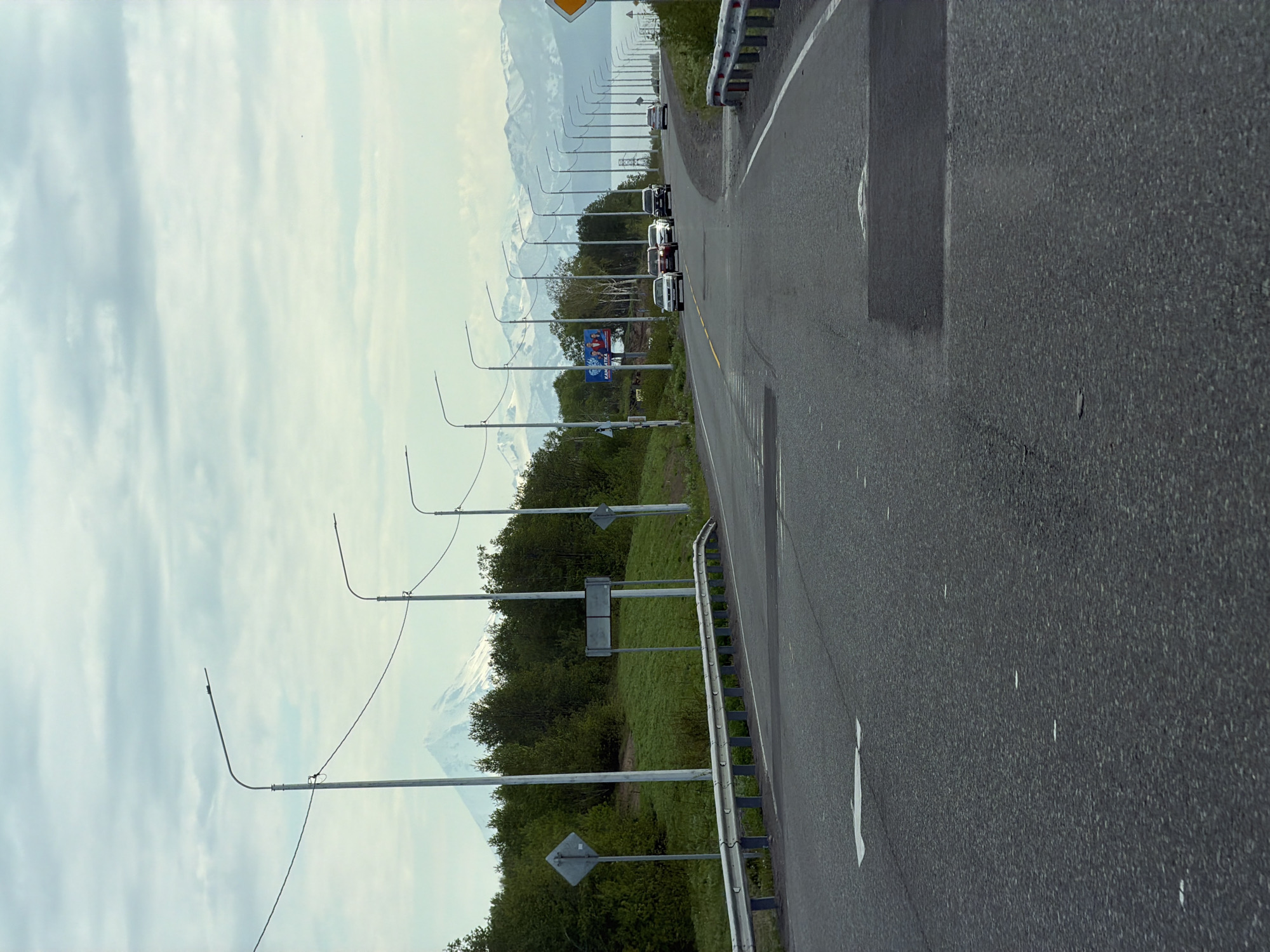 The image depicts a road in the city of Krutoberegovy, Russia, likely leading towards a mountainous or coastal area. The road is asphalted and appears to be a multi-lane highway with a guardrail on the right side. Several streetlights are visible along the side of the road, spaced at regular intervals. On the left side of the road, there is a grassy embankment with trees and a distant snow-capped mountain, possibly a volcano, in the background. A few cars are driving on the road, heading in the same direction. There are also some road signs, including a diamond-shaped sign on the left and a blank rectangular sign above it. The sky is cloudy with a hint of blue. The overall scene suggests a scenic route with natural elements and infrastructure.
