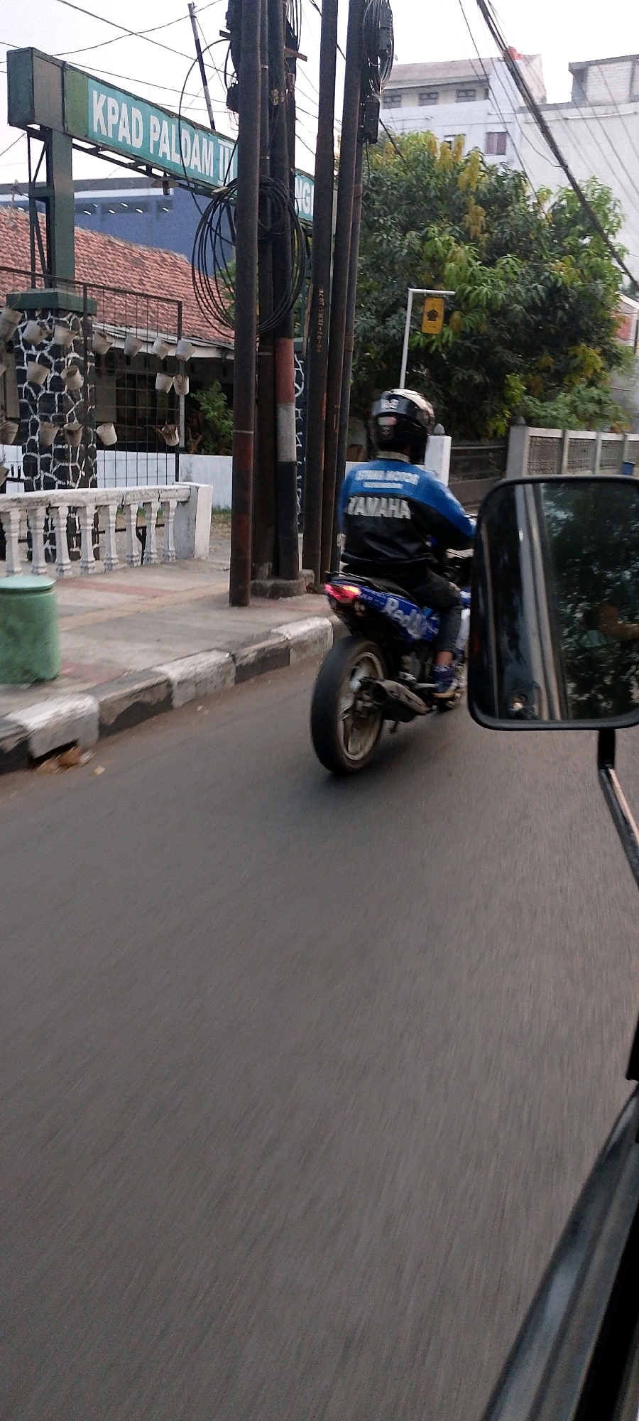 The image captures a street scene in Bandung City, Indonesia, with a focus on a motorcycle rider. The rider, wearing a black and blue jacket with "YAMAHA" prominently displayed, is seen from behind. They are on a motorcycle with a red and blue decal on the rear fender. The motorcycle is in motion on an asphalt road, indicated by motion blur. To the left, a sidewalk with a white railing and a green trash can is visible. In the background, a sign reads "KPAD PALDAM III" and a building with a red tile roof can be observed. Multiple utility poles with wires are also present in the scene. A side-view mirror of another vehicle, likely the one from which the photo was taken, is in the foreground on the right. The overall lighting suggests it is daytime.