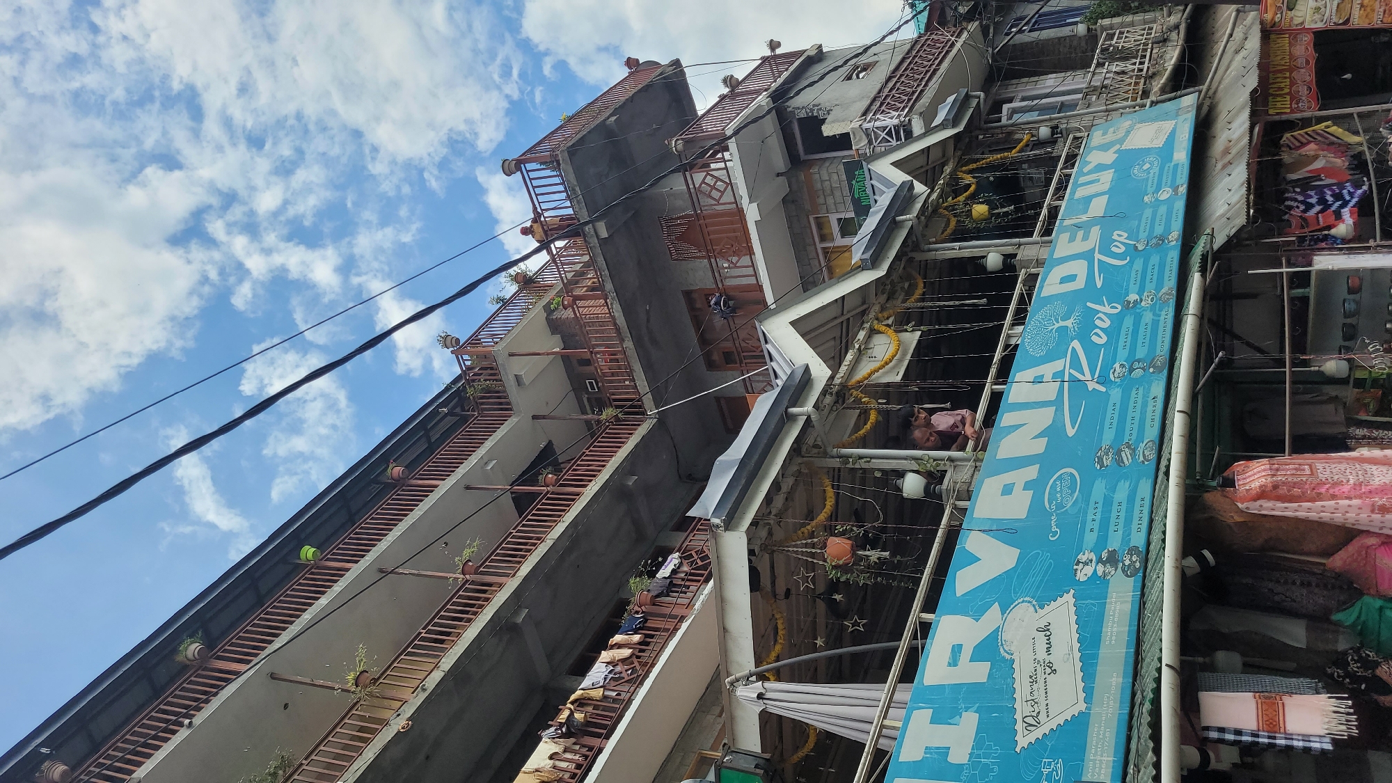 In Vashisht, India, a multi-story building is captured from a low-angle perspective, with a bright blue sky and scattered clouds above. The building features multiple levels with balconies, each adorned with wooden railings and potted plants. Laundry hangs from lines on one of the balconies. Below the balconies, a covered area with decorative yellow garlands and lights appears to be a restaurant or cafe. A prominent blue sign for "NIRVANA DELUXE Roof Top" is visible, listing various food categories. Two individuals, appearing to be men, are looking over the railing of the cafe area. To the left of the building, clothes are displayed for sale, hanging on racks. In the lower right corner, a sign for "THE CAFE VASHISH" is visible. Power lines crisscross the upper portion of the image.